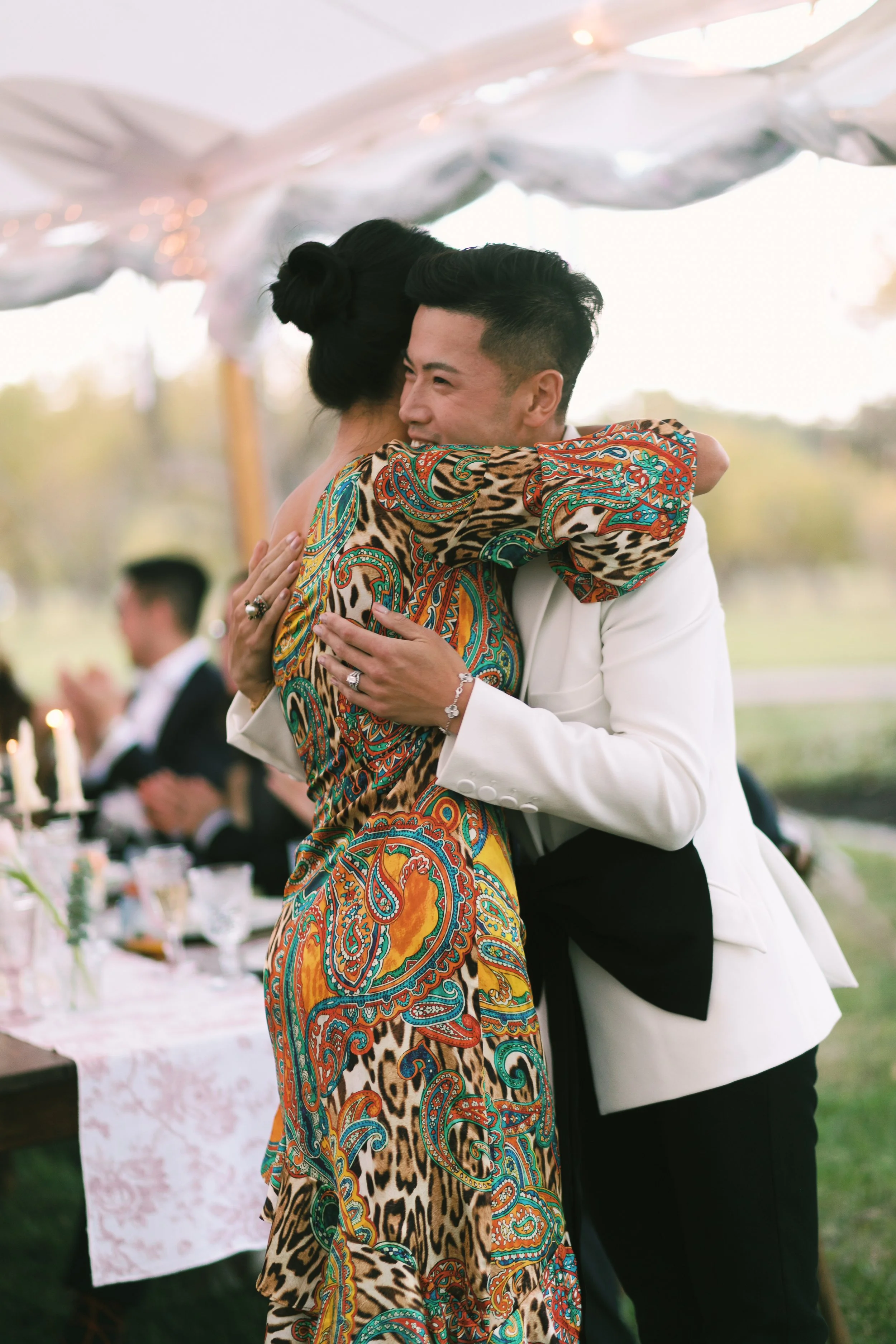 A man and woman embracing at an outdoor event, with the man wearing a white suit and the woman wearing a colorful patterned dress. Other people are seen clapping in the background.