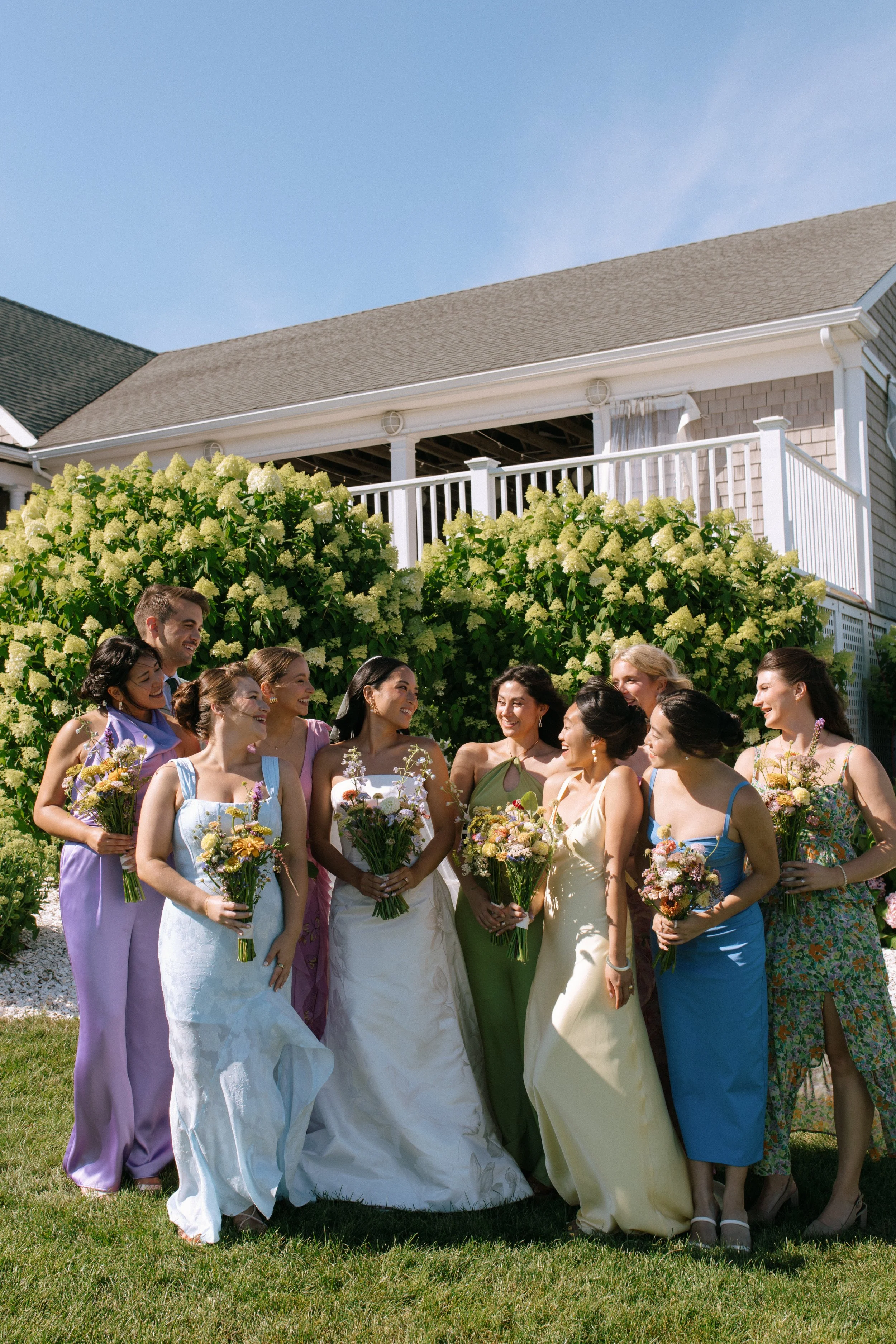 A group of women in colorful dresses holding bouquets of flowers, gathering outdoors in front of lush green bushes and a house with a balcony, during a sunny day.