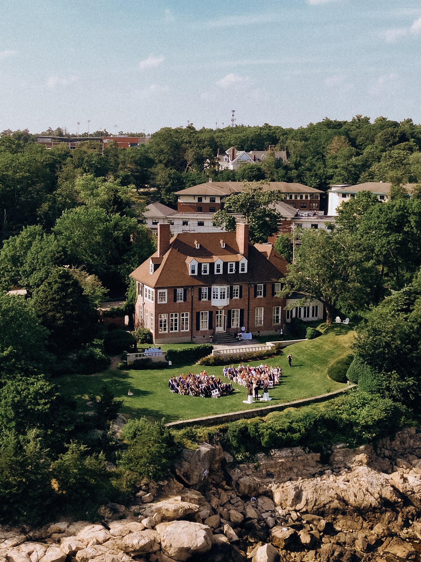 An outdoor wedding reception in front of a large brick house with a lawn and surrounded by trees, on a sunny day.