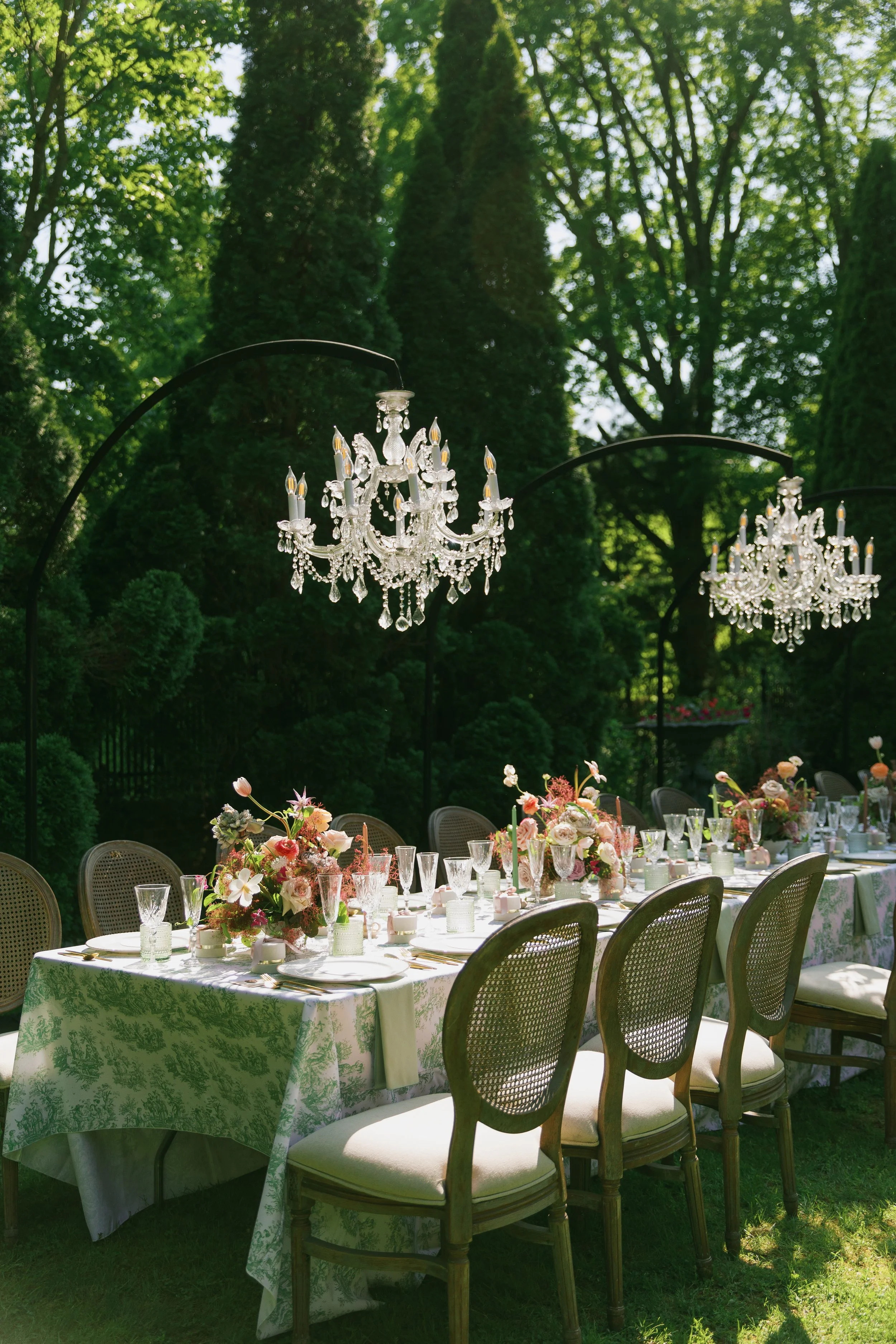 Elegant outdoor dining table set for a formal event with floral centerpieces, crystal glassware, and chandeliers hanging above in a lush garden setting.