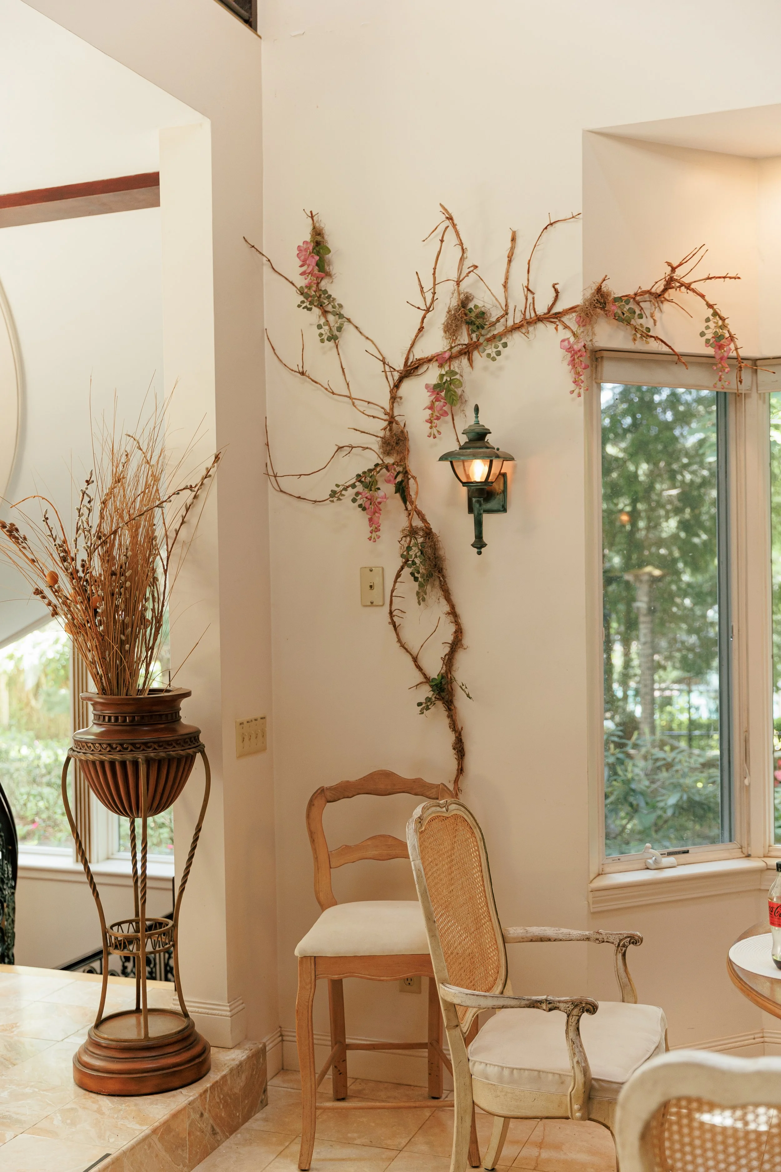 Decorative wall with bare vine branch and pink flowers, vintage wall sconce with light, and two chairs near a window with greenery outside.