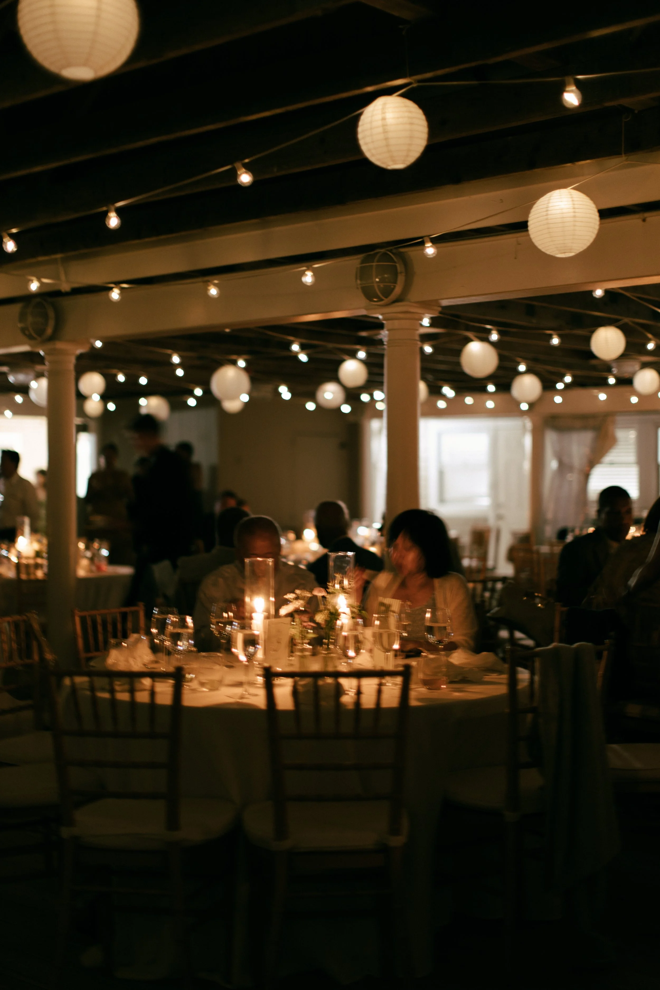 Indoor dining area with round tables, candlelit centerpieces, and paper lanterns hanging from the ceiling, filled with guests enjoying a meal.