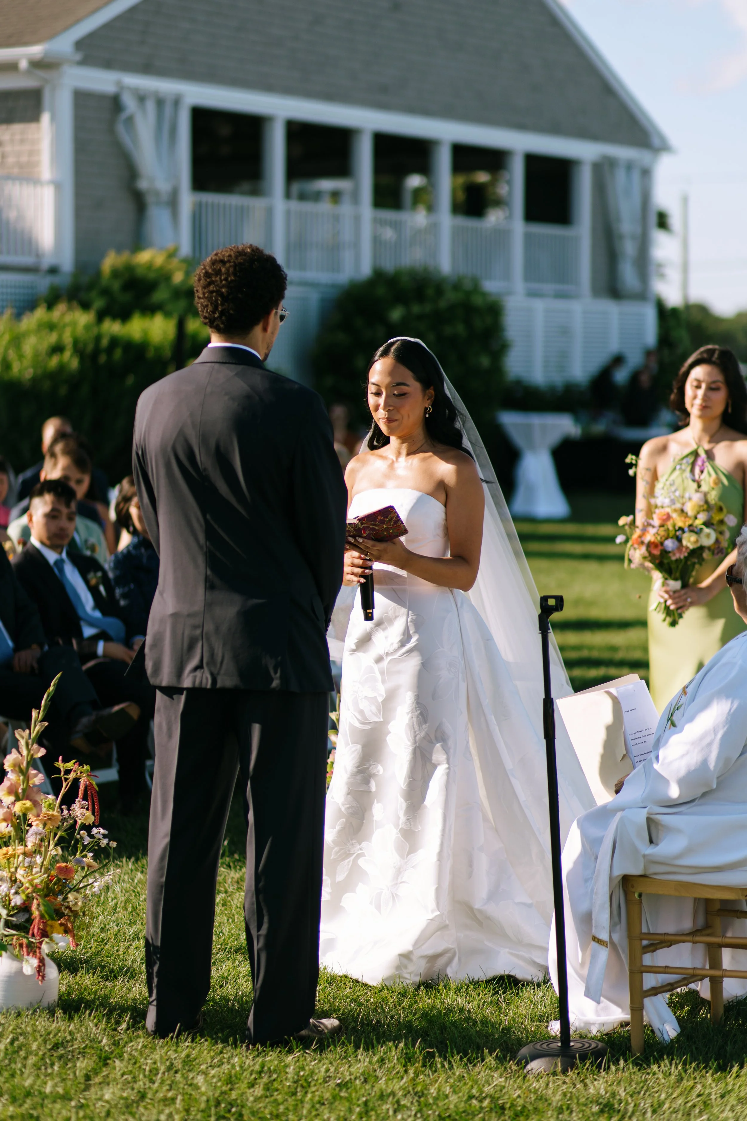 A bride and groom exchanging vows during a wedding ceremony outdoors, with guests seated in the background and a woman in a green dress holding a bouquet standing nearby.