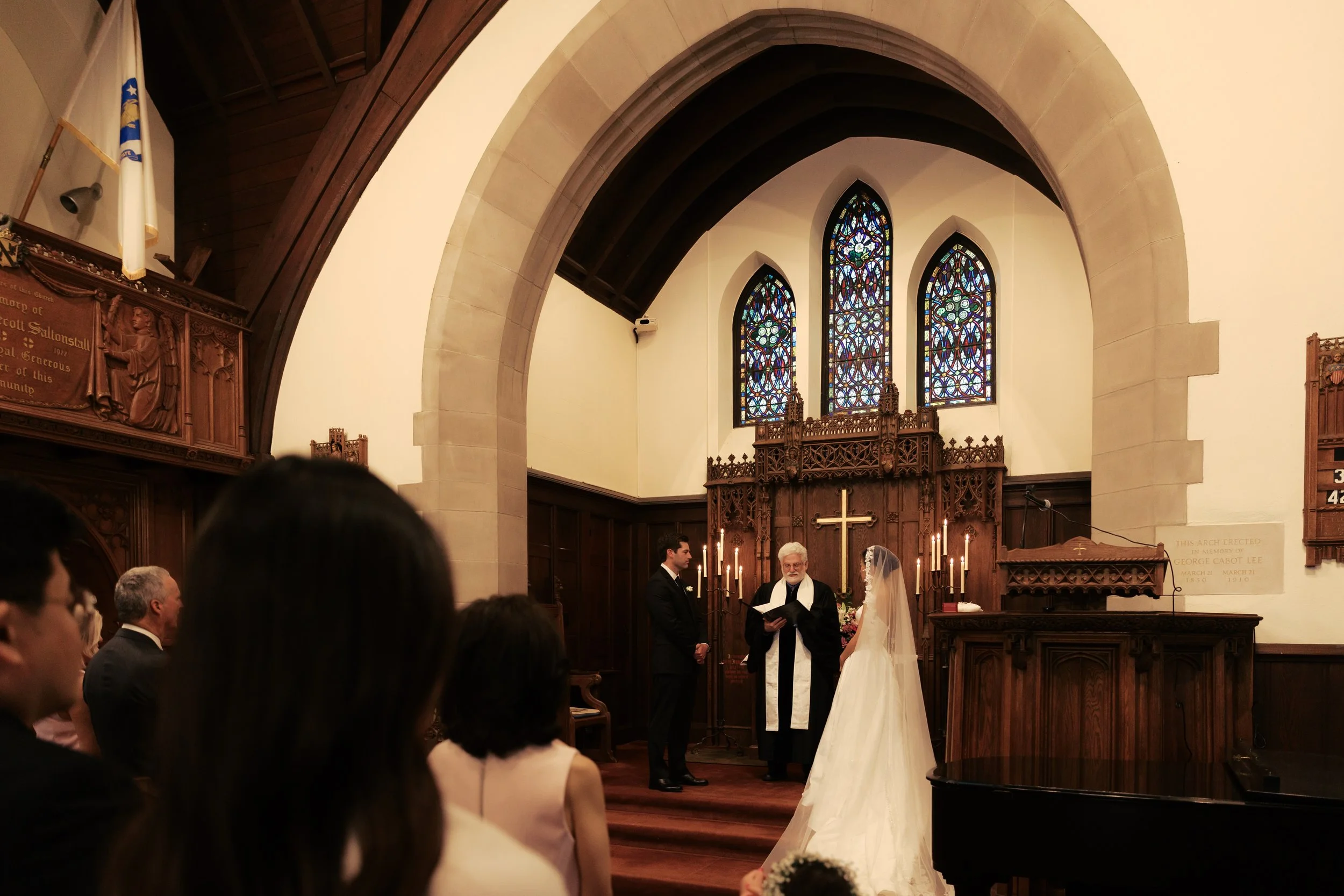 A wedding ceremony inside a church with stained glass windows. The bride in a white dress and veil stands before a minister. The groom, in a dark suit, faces her. Guests are seated, watching the couple. The wooden altar has a gold cross and lit candl