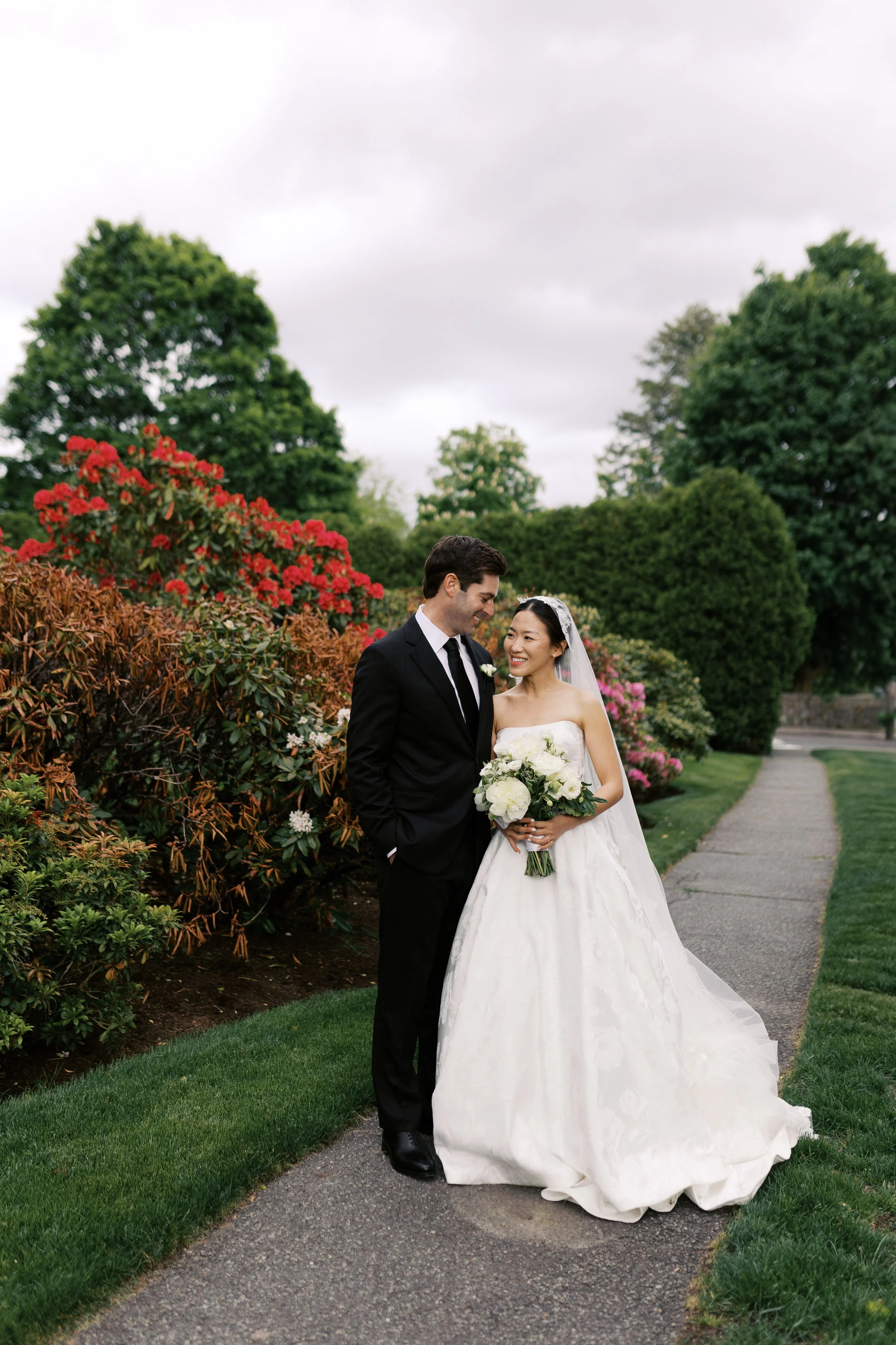 A bride and groom standing on a garden path, smiling and looking at each other, surrounded by green trees and blooming bushes.