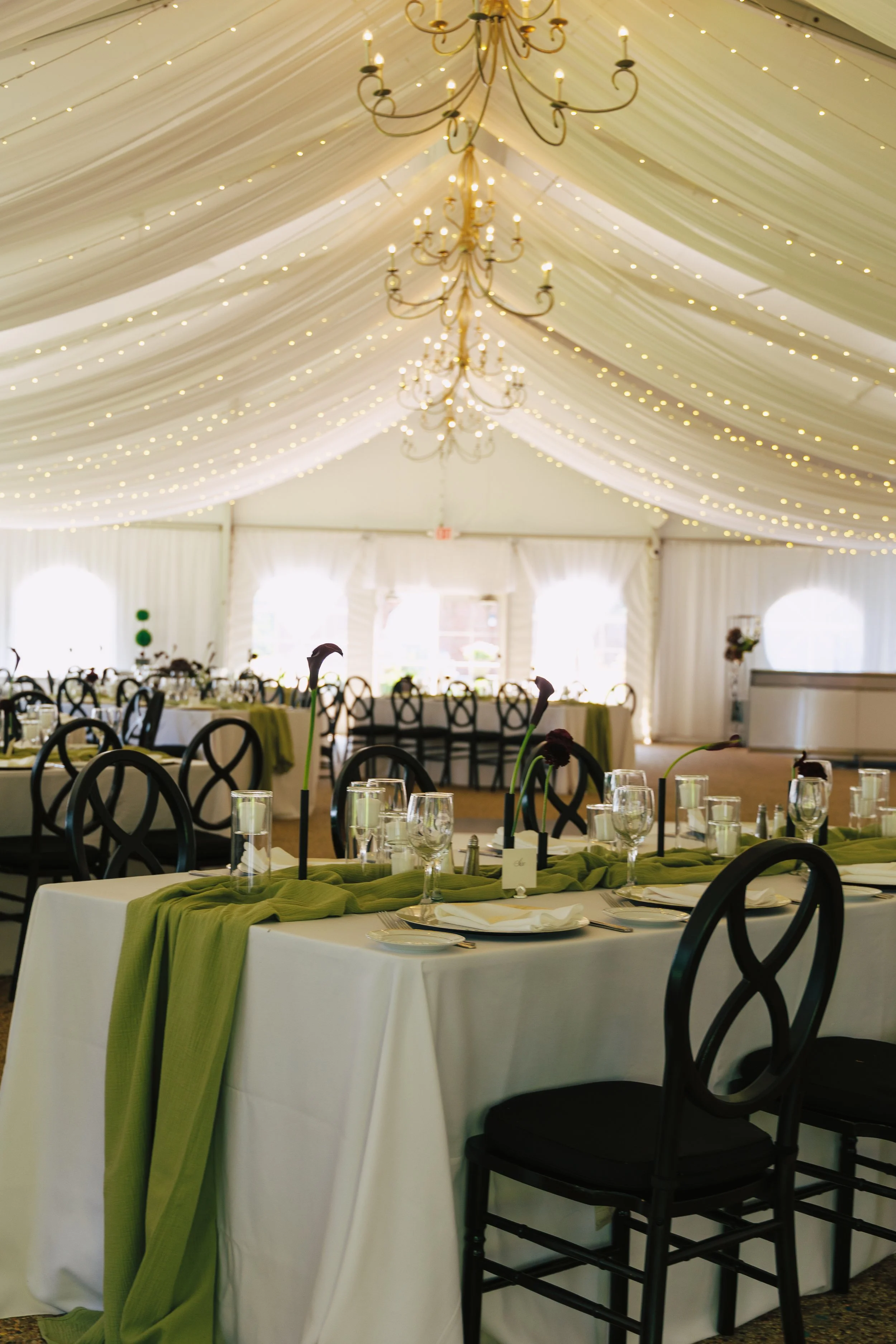 Elegant event tent with draped white fabric ceiling, string lights, and chandeliers. Indoor tables are decorated with green runners, tall calla lily centerpieces, and set with glassware, plates, and utensils.