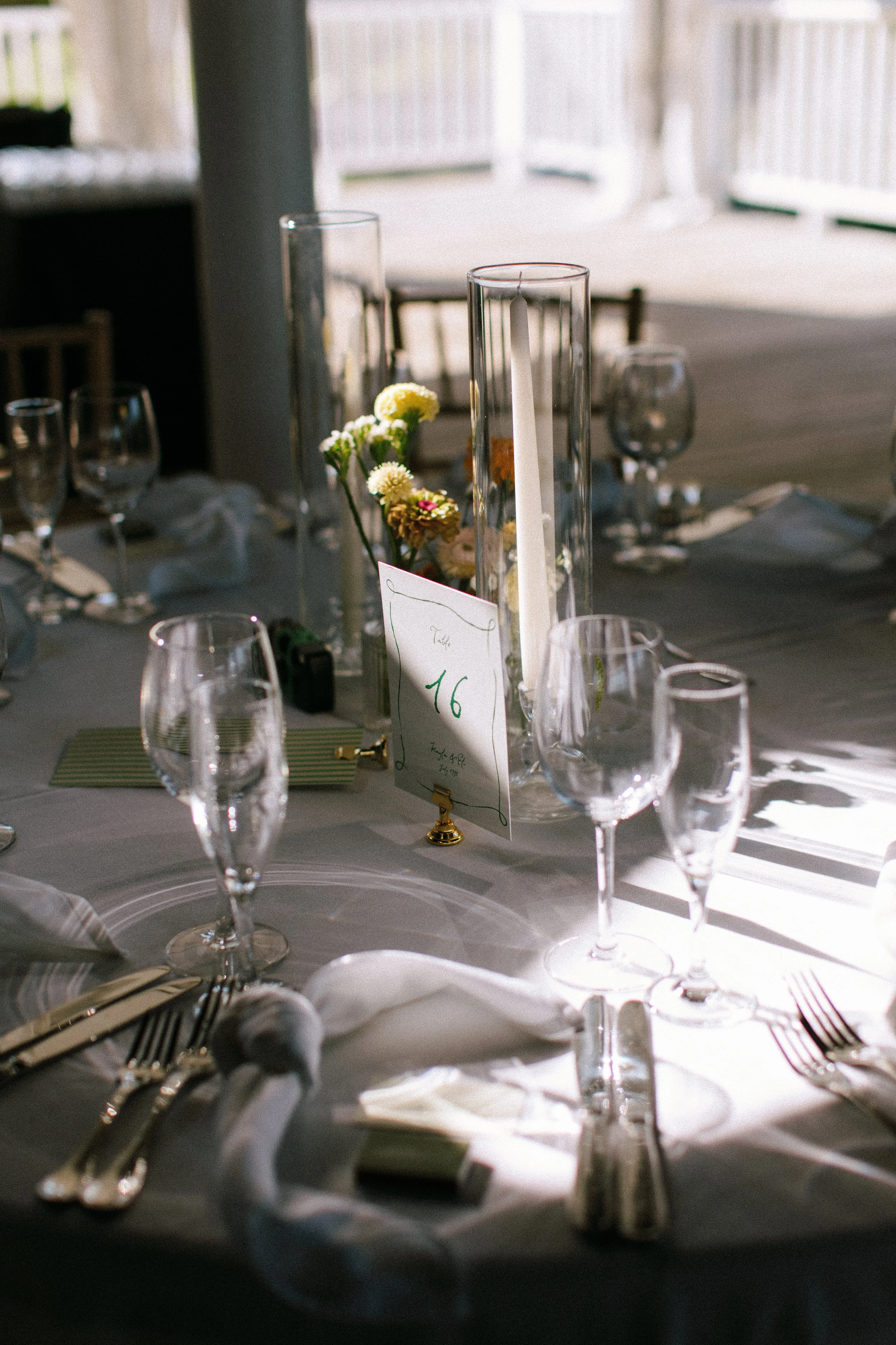 A formal dining table set with glassware, silverware, napkins, a table number card, and a floral centerpiece featuring white and yellow flowers.
