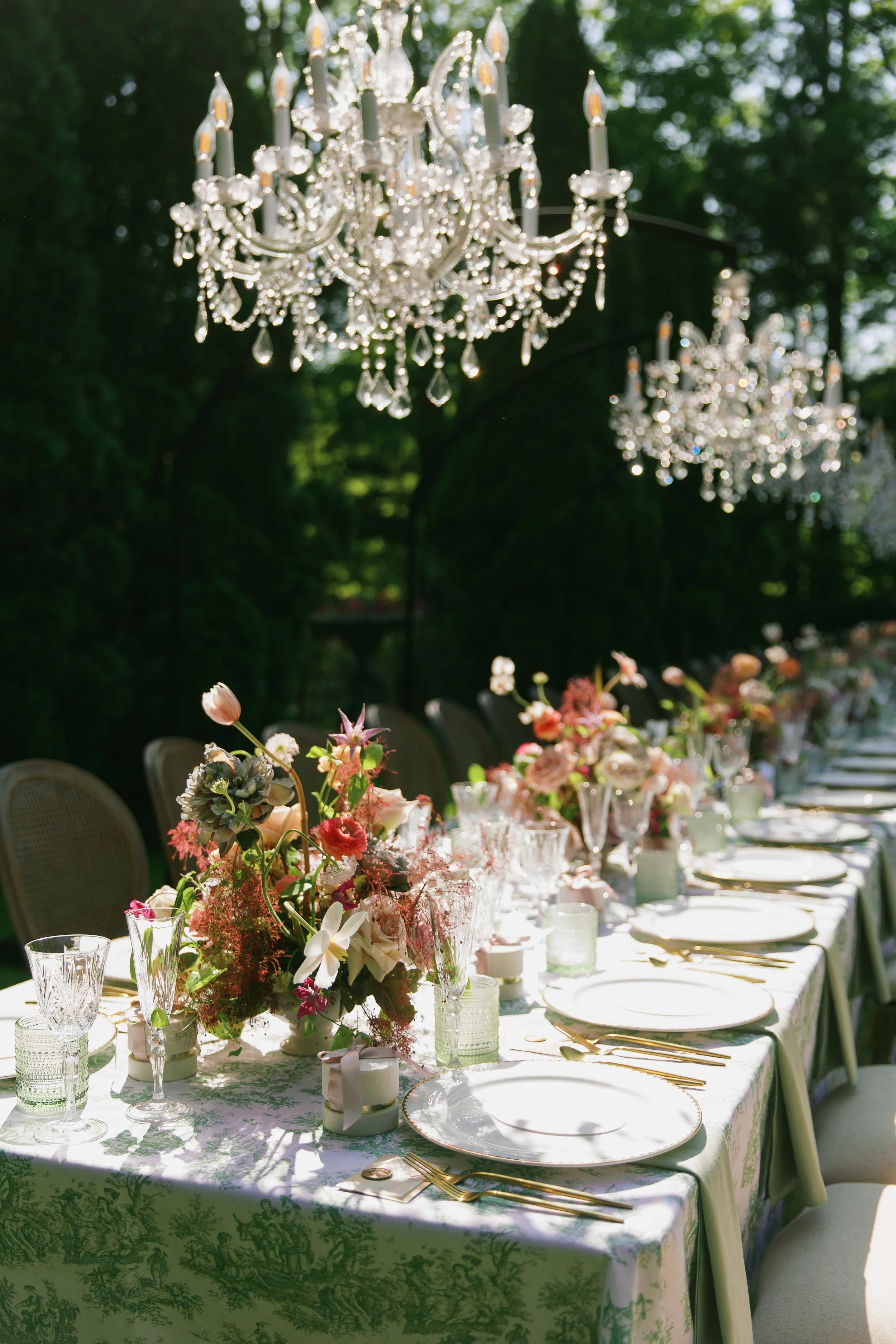 A long dining table set outdoors with floral centerpieces, white plates, and gold cutlery, illuminated by two large chandeliers hanging above on a sunny day.