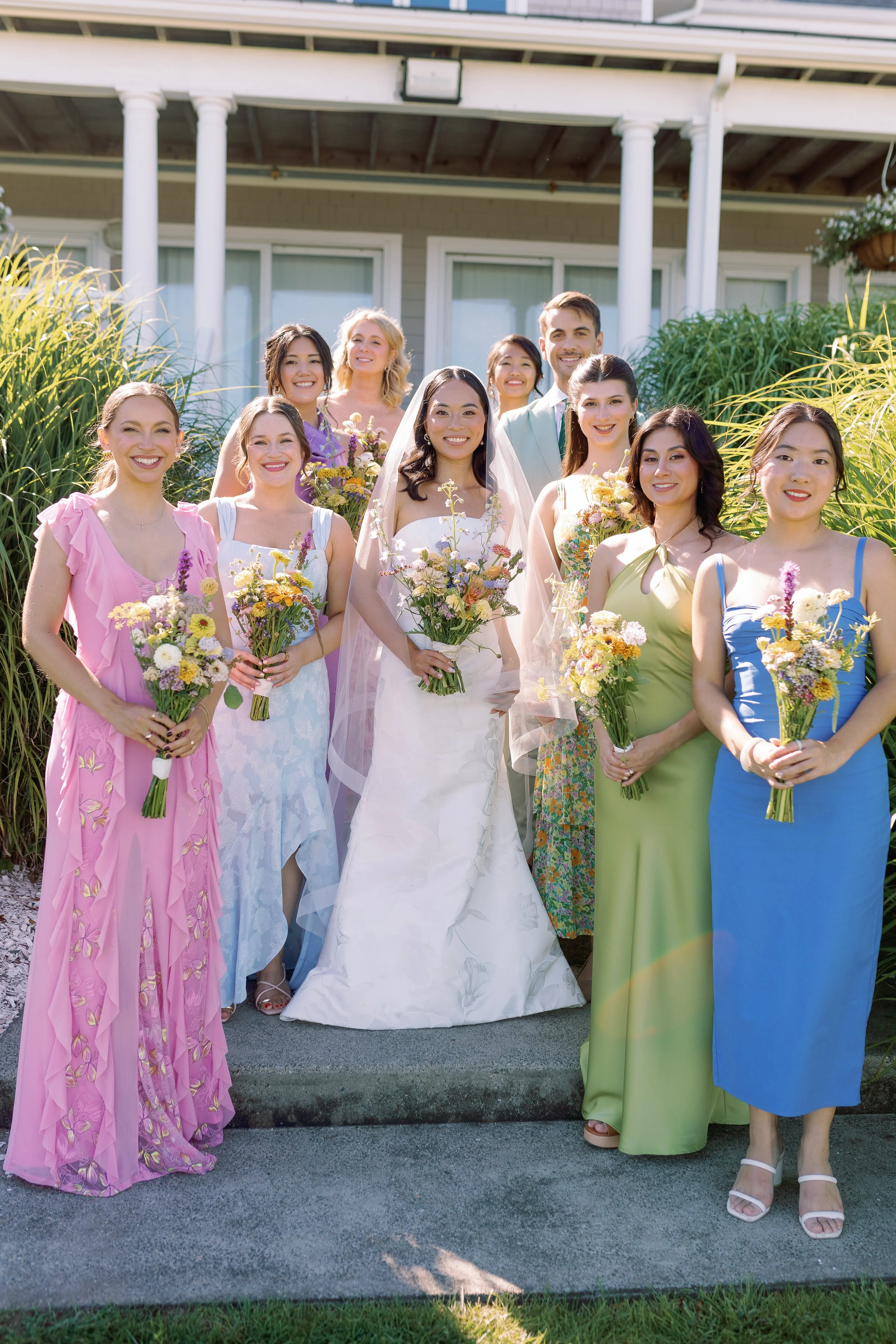 A wedding group photo with the bride in a white wedding gown and veil, surrounded by bridesmaids in colorful dresses, all holding bouquets of flowers, posing outdoors in front of a house with columns and greenery.