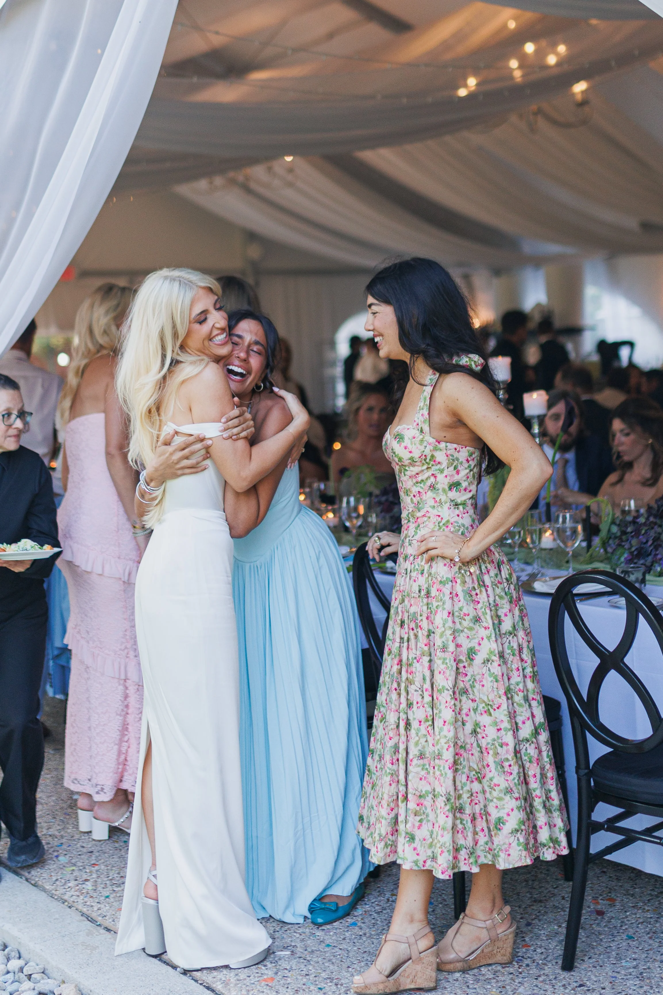 Four women celebrating at a social event under a decorated tent, with tables set with candles, flowers, and glassware.