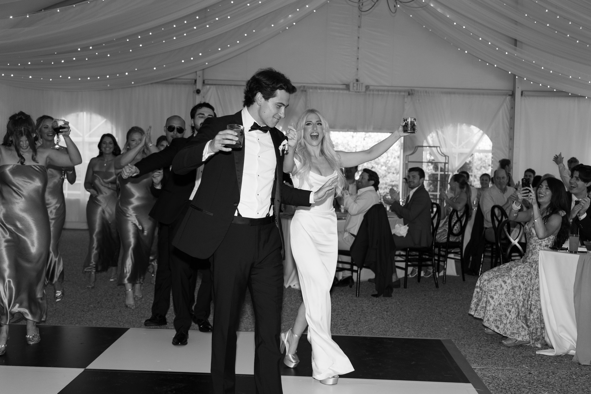 Black-and-white photo of a wedding reception, with a groom and bride leading a dance or parade, surrounded by guests clapping and cheering in a decorated banquet hall.
