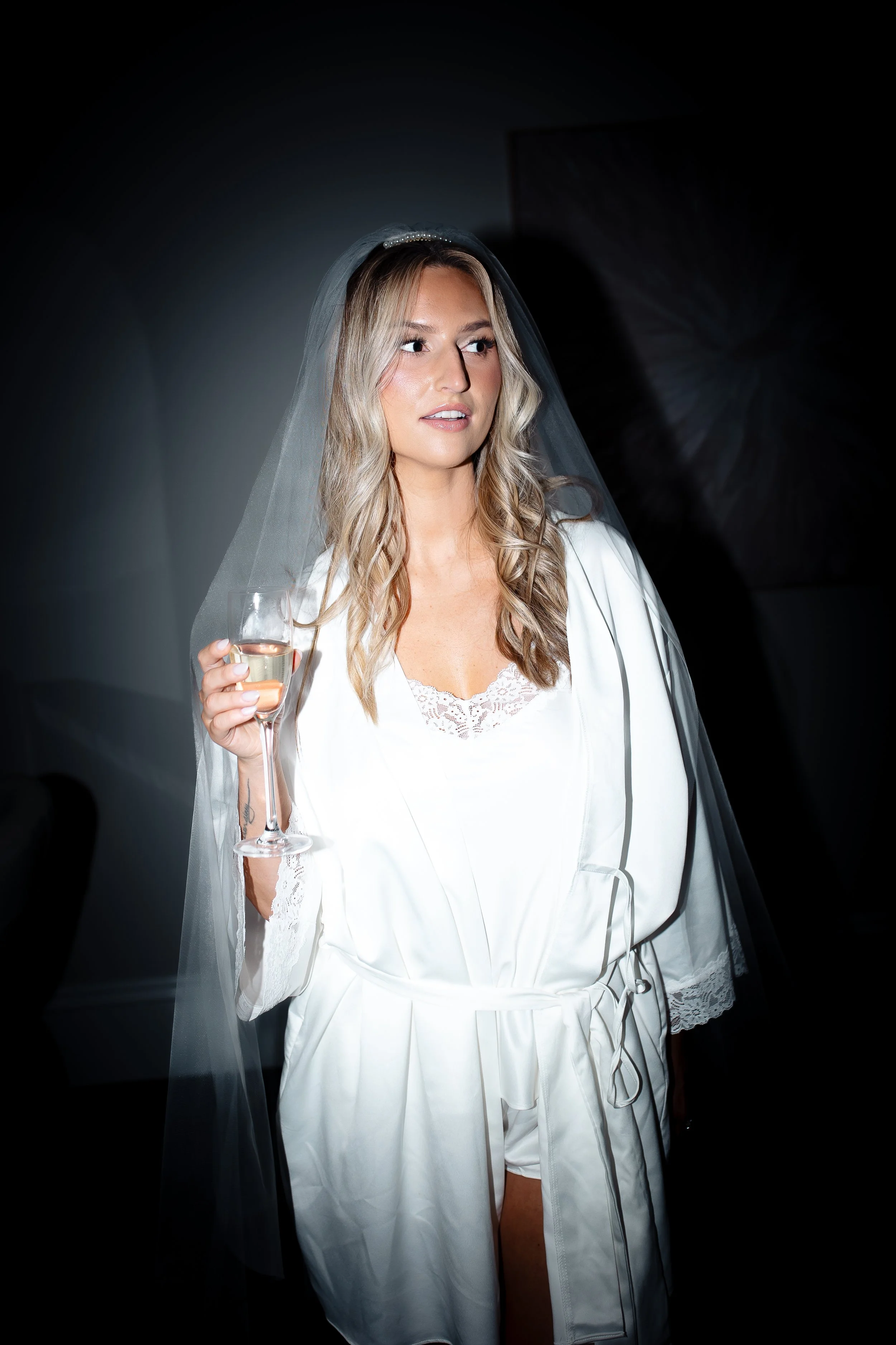 A woman with long blonde hair wearing a white robe and veil holding a glass of champagne.