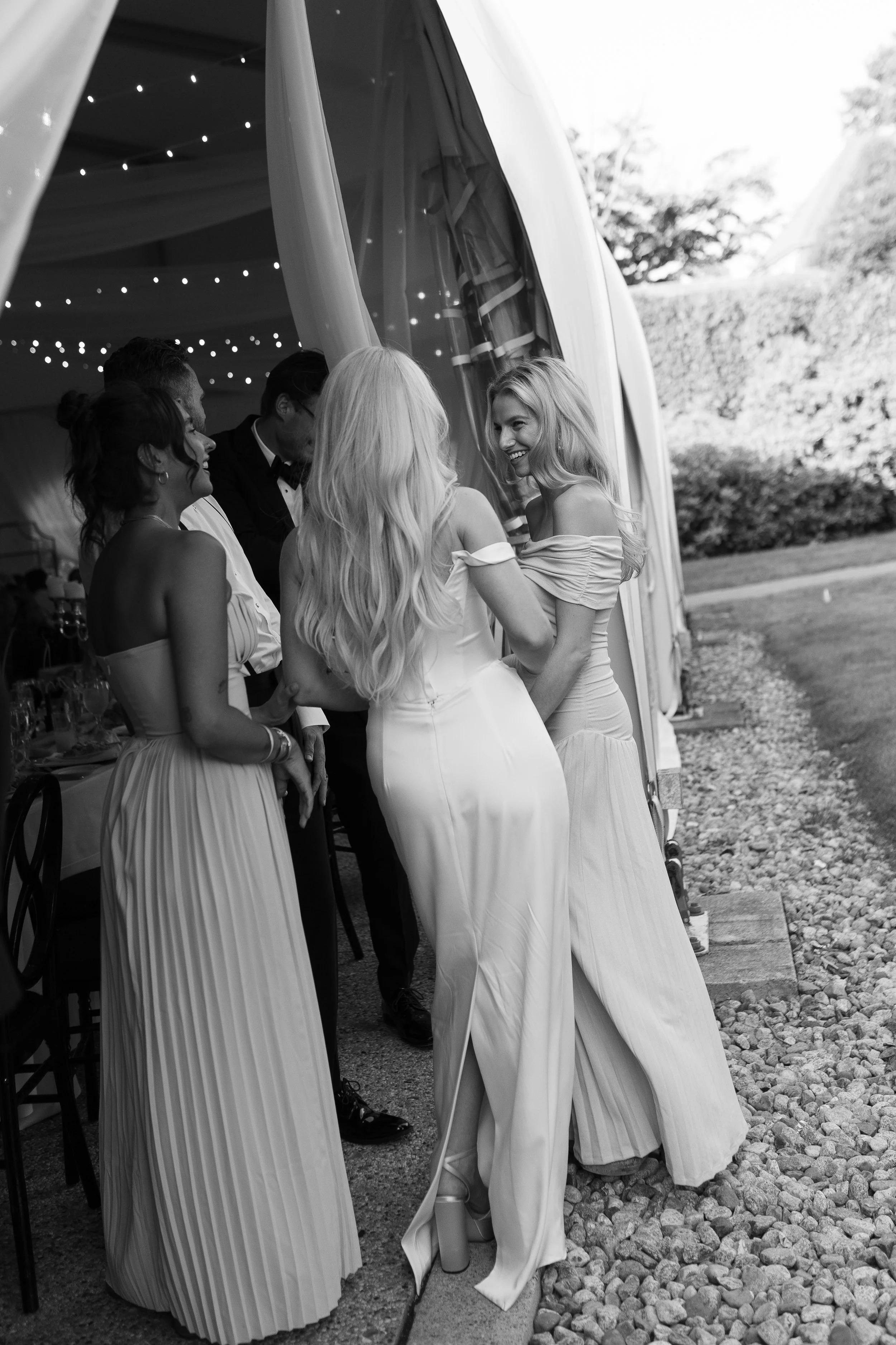 Women in elegant dresses smiling and chatting at an outdoor event, possibly a wedding, under a tent with string lights.