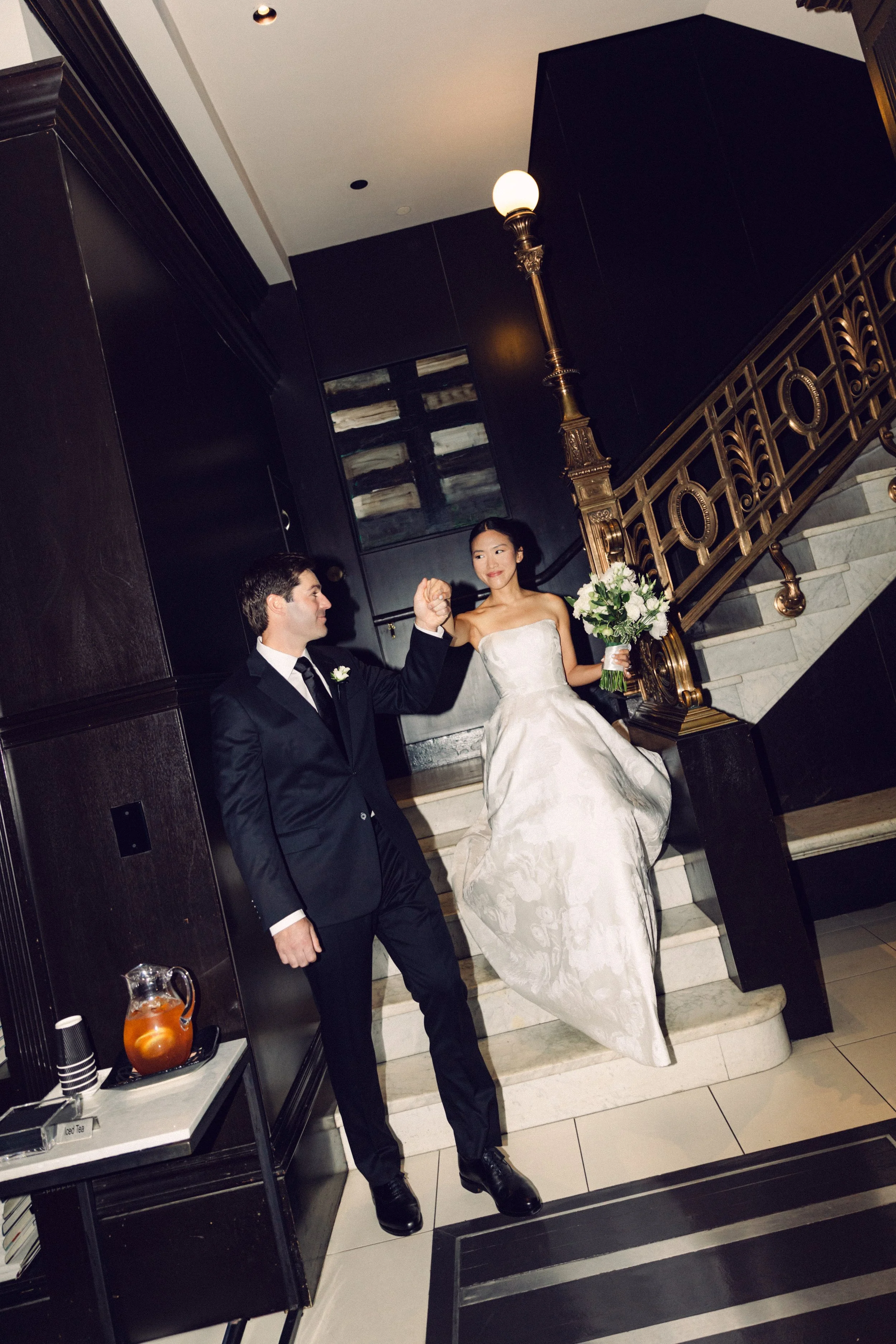A bride in a white strapless wedding gown holding a bouquet of white flowers stands on a staircase, holding hands with a groom in a black tuxedo. The setting is an elegant indoor space with dark walls, a gold railing, and a white marble staircase.