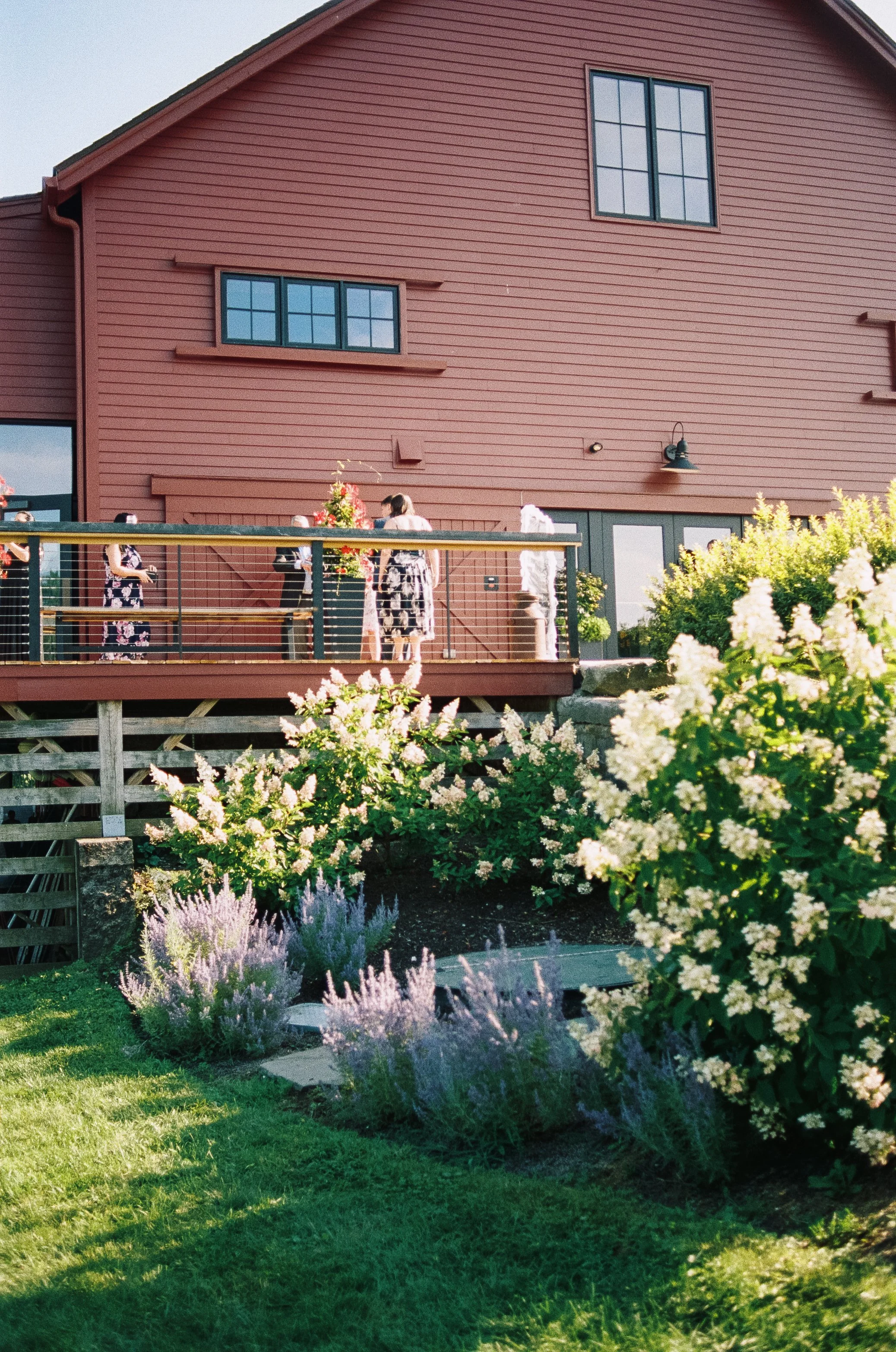 People gathered on a wooden deck outside a red barn-style building with floral decorations and green plants.