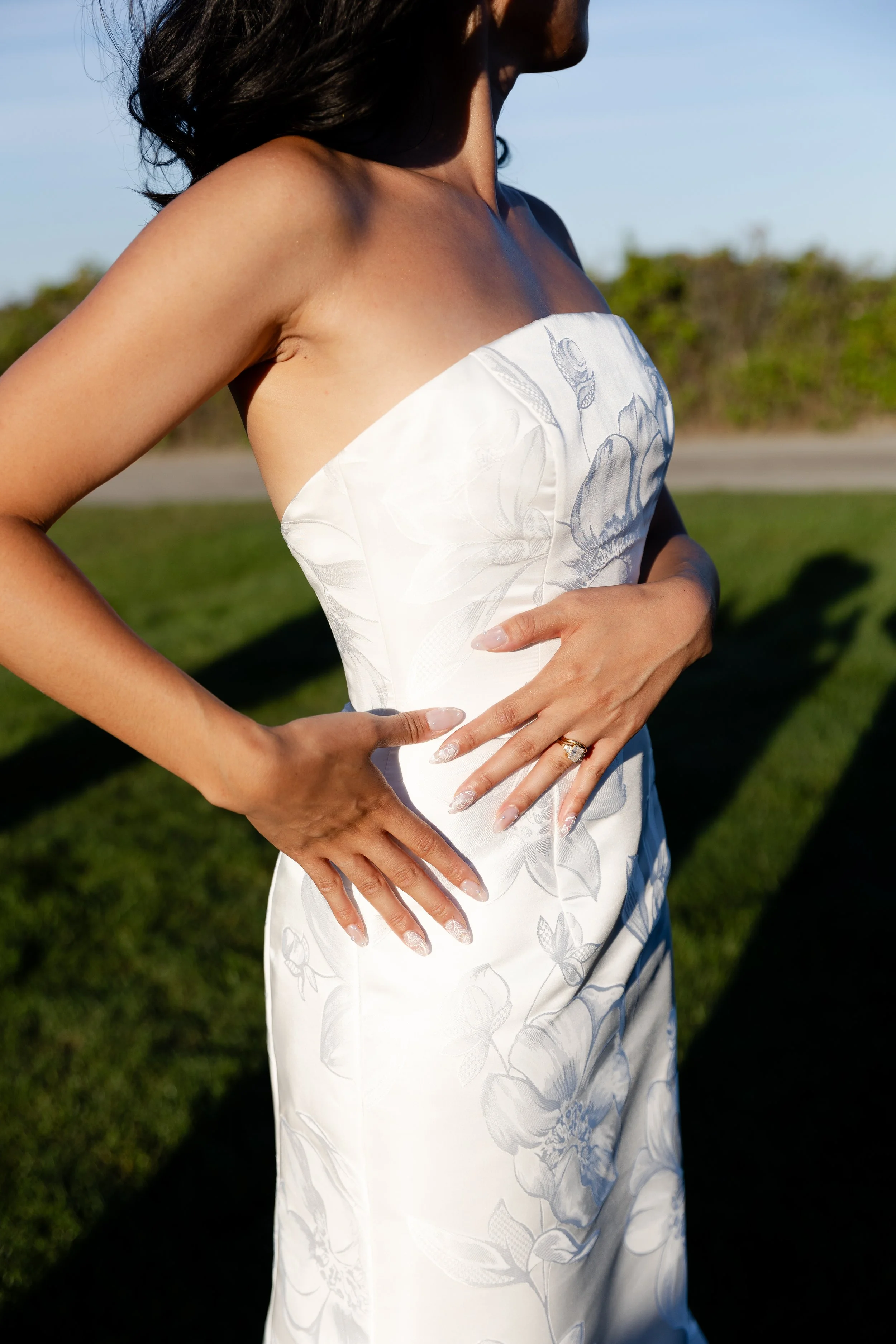 A woman in a strapless white floral dress standing outdoors in the sunlight with her hands resting on her midsection, showing a gold ring on her left hand.