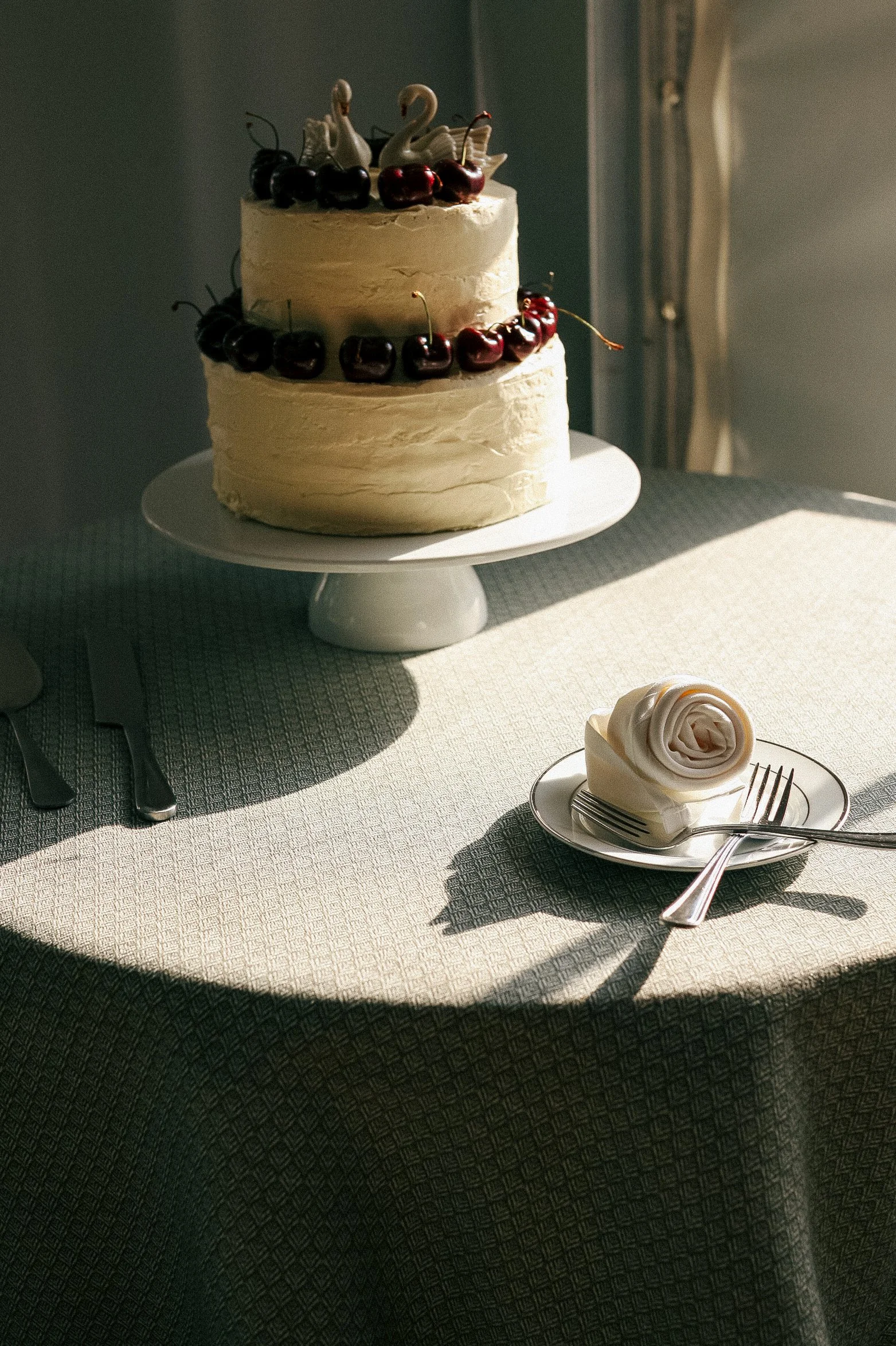 A two-tier white frosted cake decorated with cherries and swan-shaped figurines on top, placed on a white cake stand, with a slice of cake with a rose-shaped frosting decoration on a plate and two forks beside it on a table with a textured tablecloth