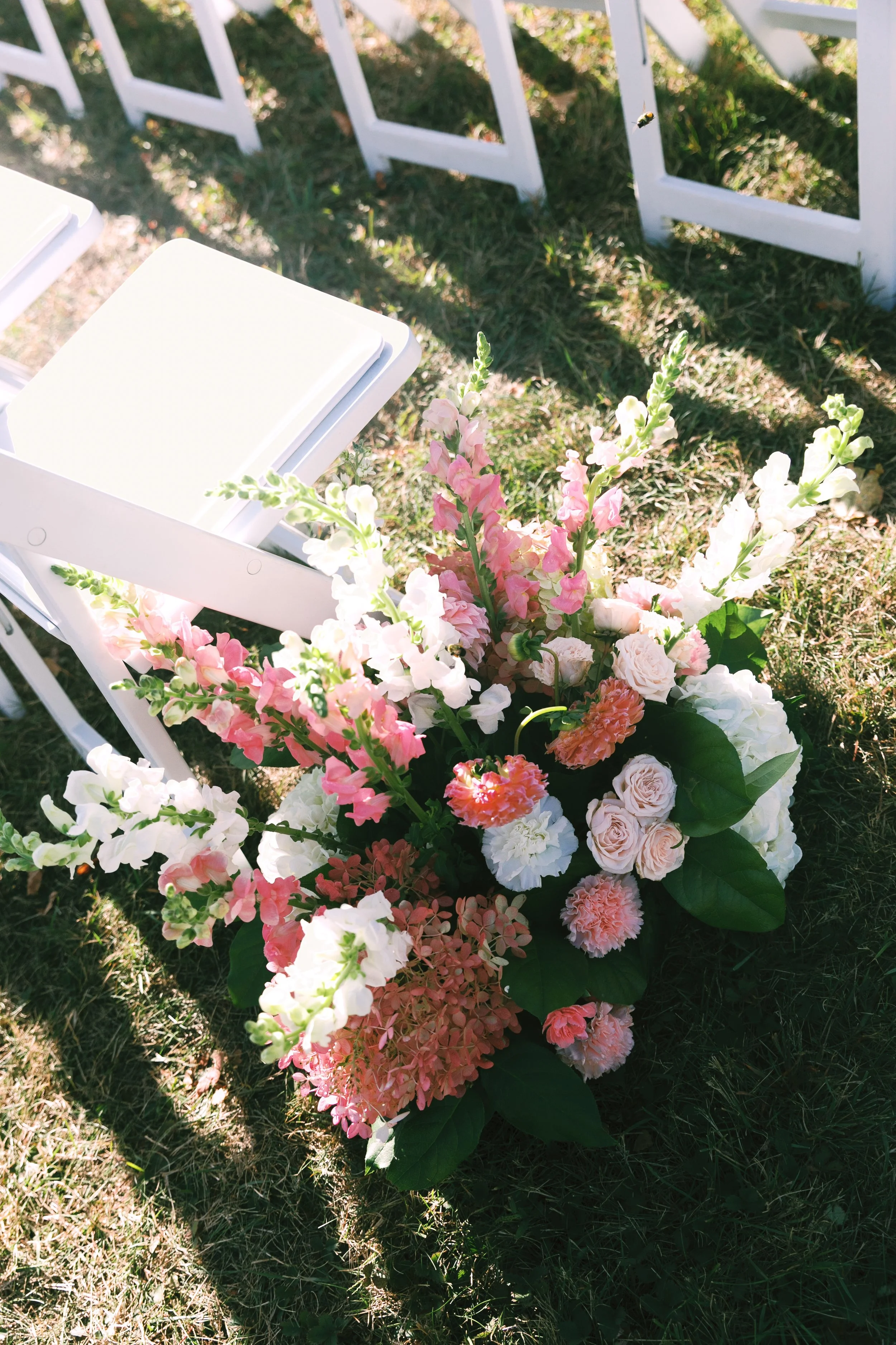 A floral arrangement with pink, white, and peach flowers, placed on the ground next to white chairs arranged outdoors on grass, illuminated by sunlight.