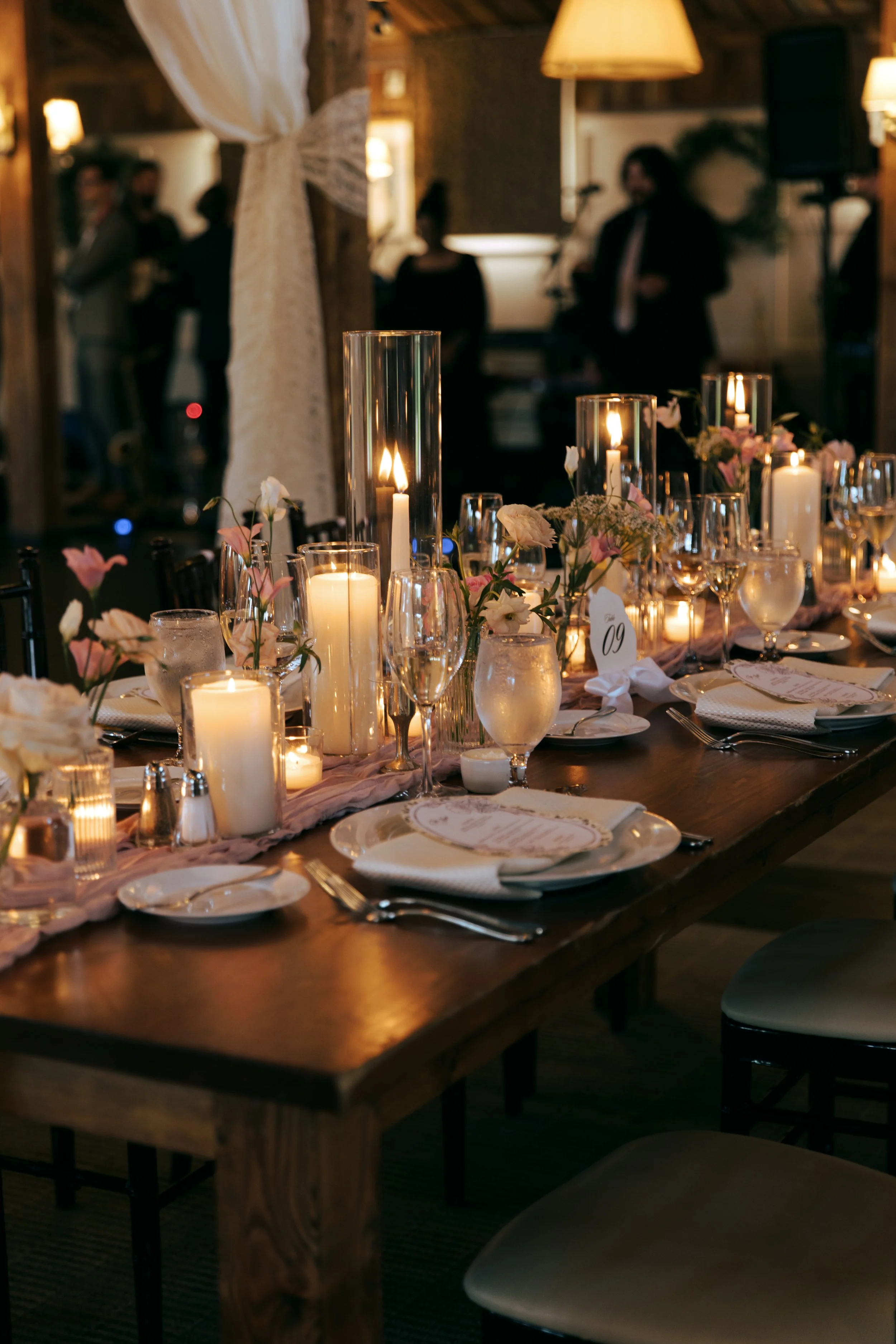 A decorated dining table with lit candles, flower arrangements, and place settings in a dimly lit event space, likely for a wedding or formal occasion, with guests in background.
