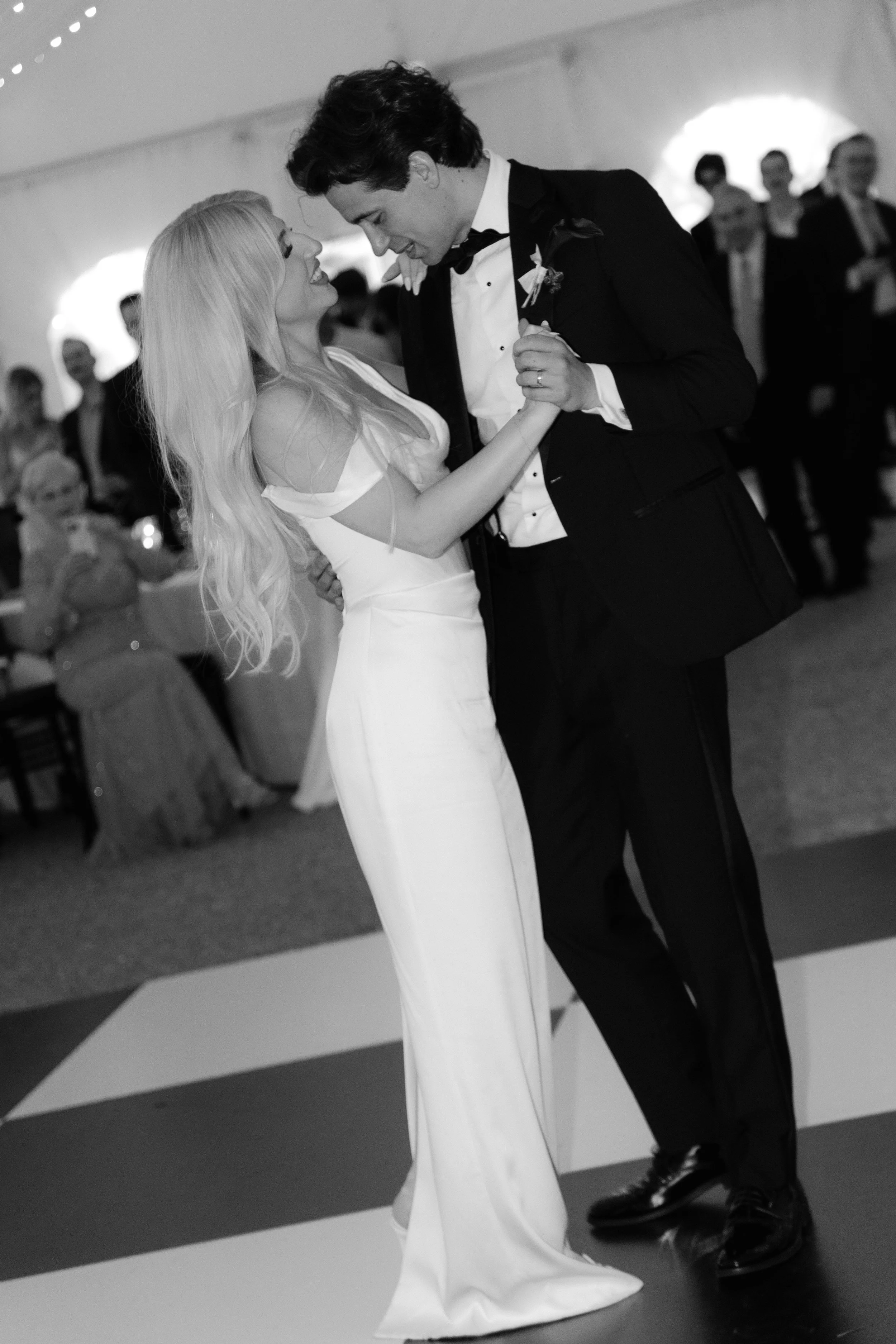 A couple dancing closely at a wedding reception, with guests in the background inside a decorated tent.