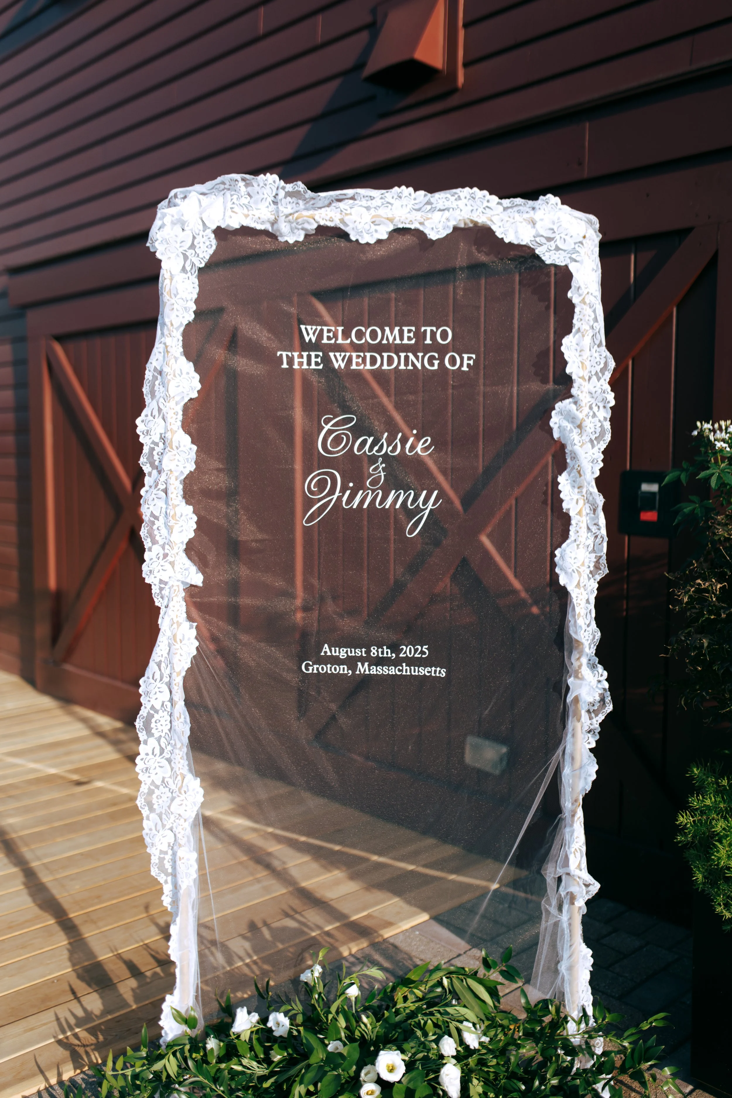 Wedding welcome sign with lace border on a transparent background, displaying details of Cassie and Jimmy's wedding on August 8th, 2025, in Groton, Massachusetts, placed outdoors on a wooden deck.