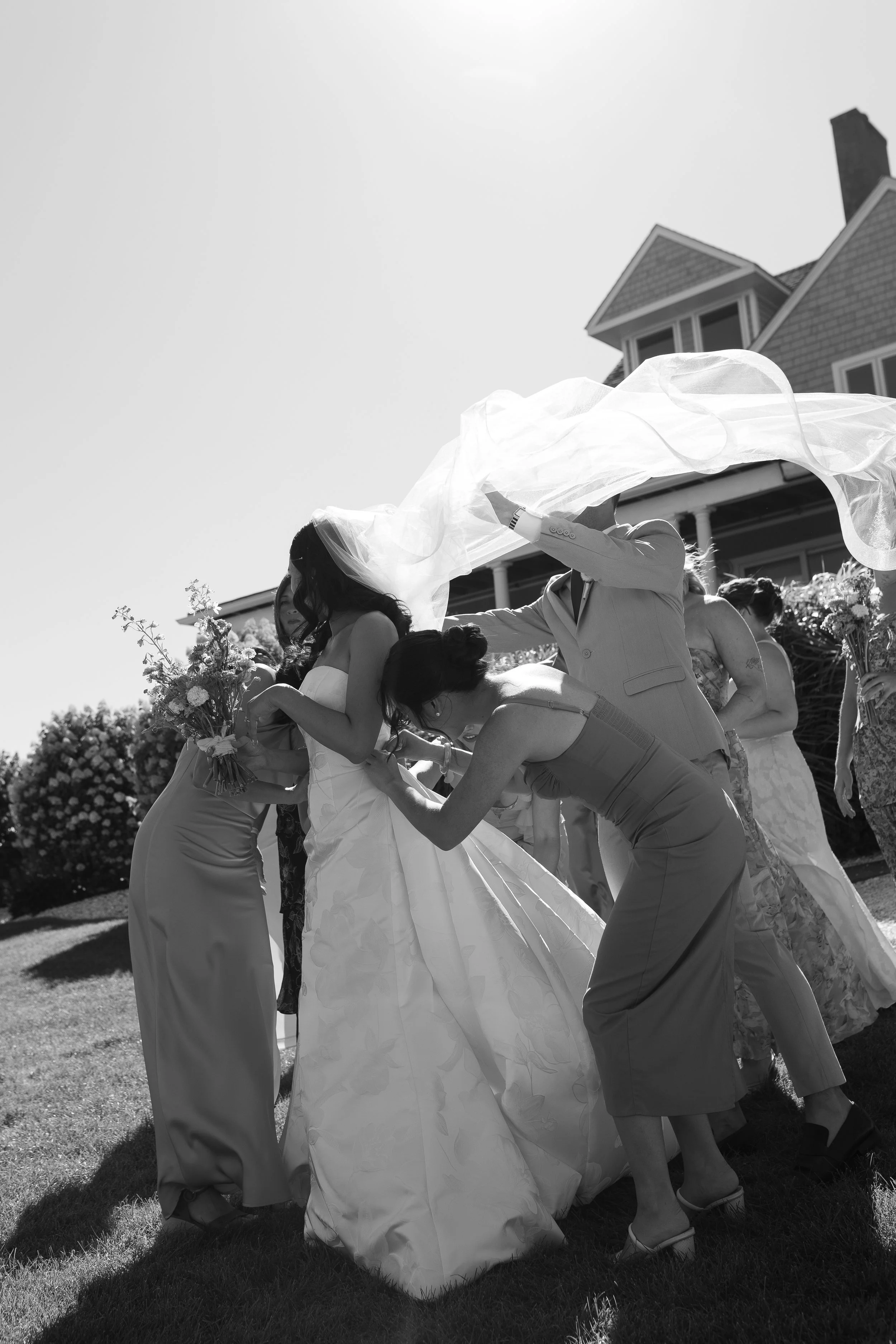 A black and white photo of a bride being assisted by friends during a wedding, with several women wearing dresses and holding flowers, outdoors near a house.