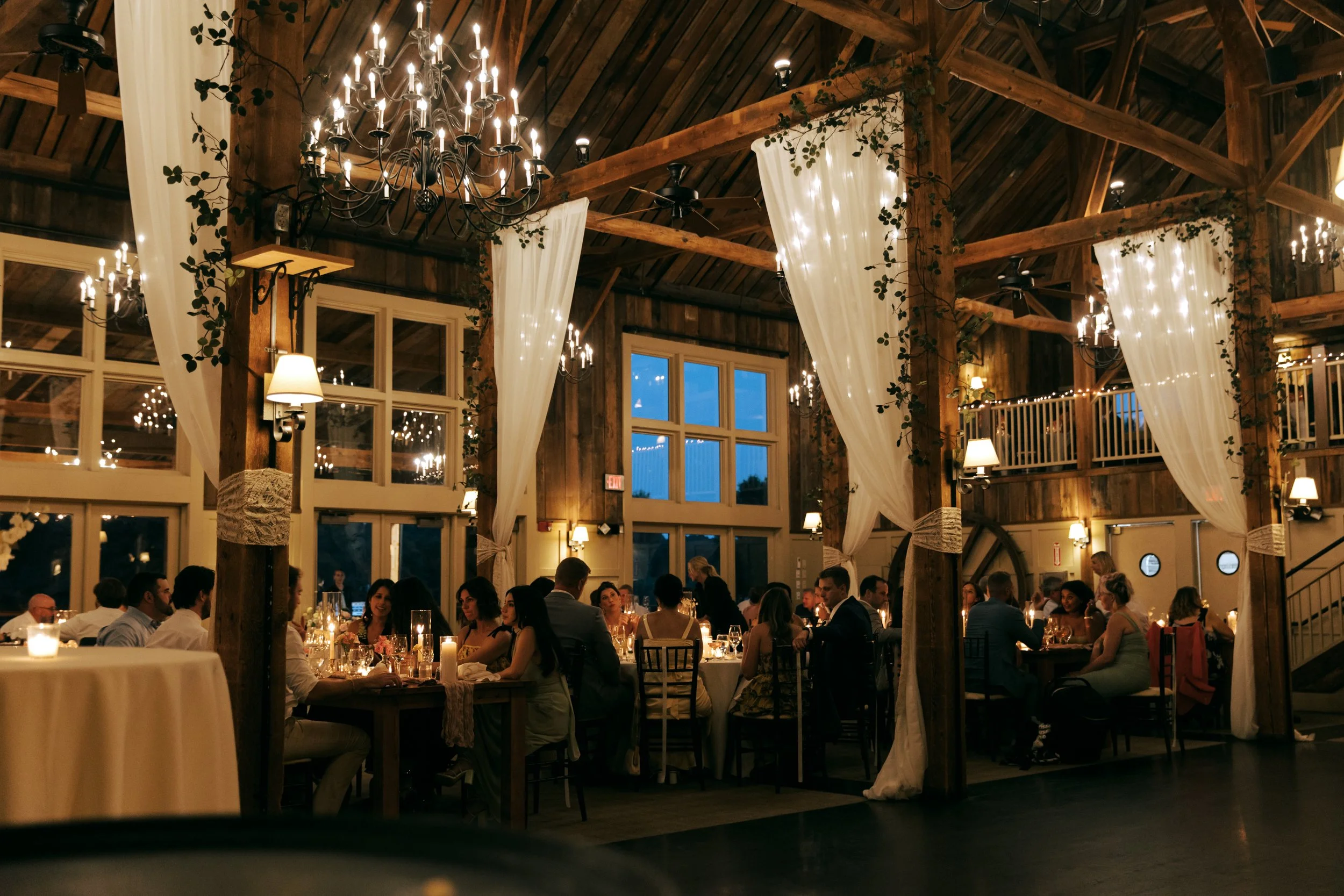 An indoor rustic Barn at Gibbet Hill Groton wedding reception with wooden beams, chandeliers, string lights, and white curtains, filled with guests seated at tables.