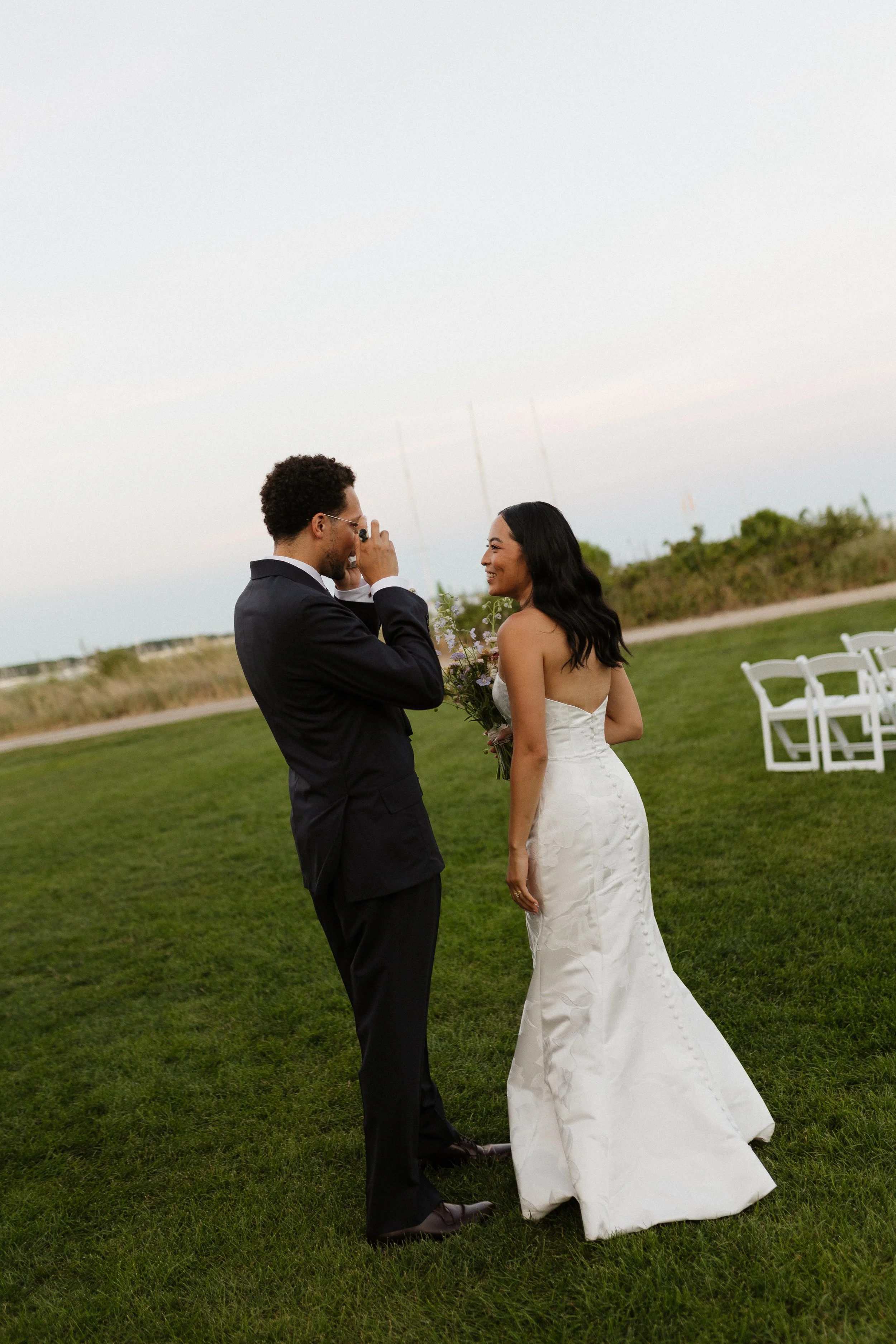A couple outdoors at a wedding, the man in a black tuxedo taking a photo of the woman in a white wedding dress holding a bouquet of flowers, on a grassy field with white chairs in the background.