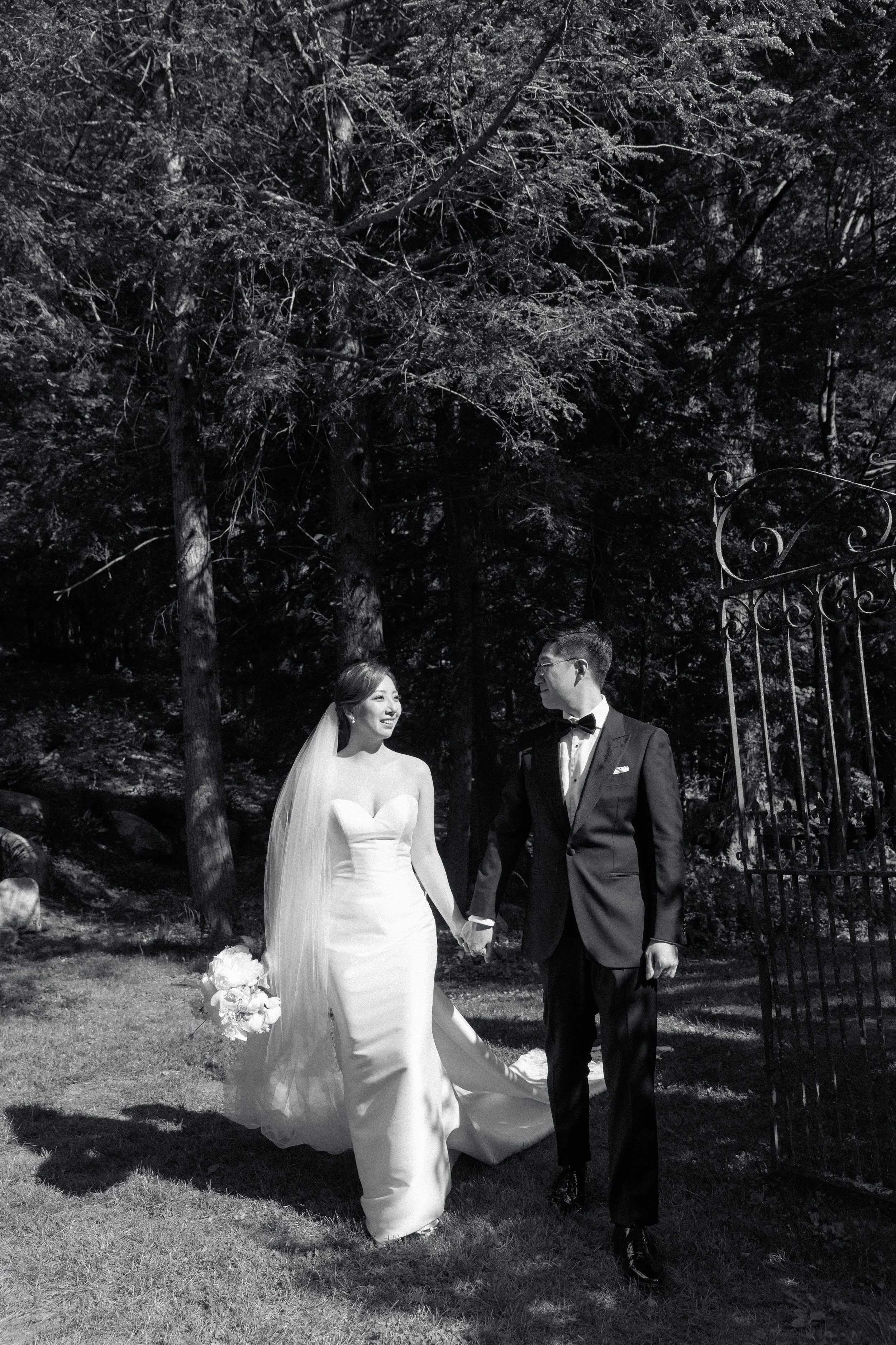 A bride and groom holding hands in a wooded outdoor area during their wedding, with the bride in a strapless wedding gown and veil, and the groom in a tuxedo, near a decorative metal gate.