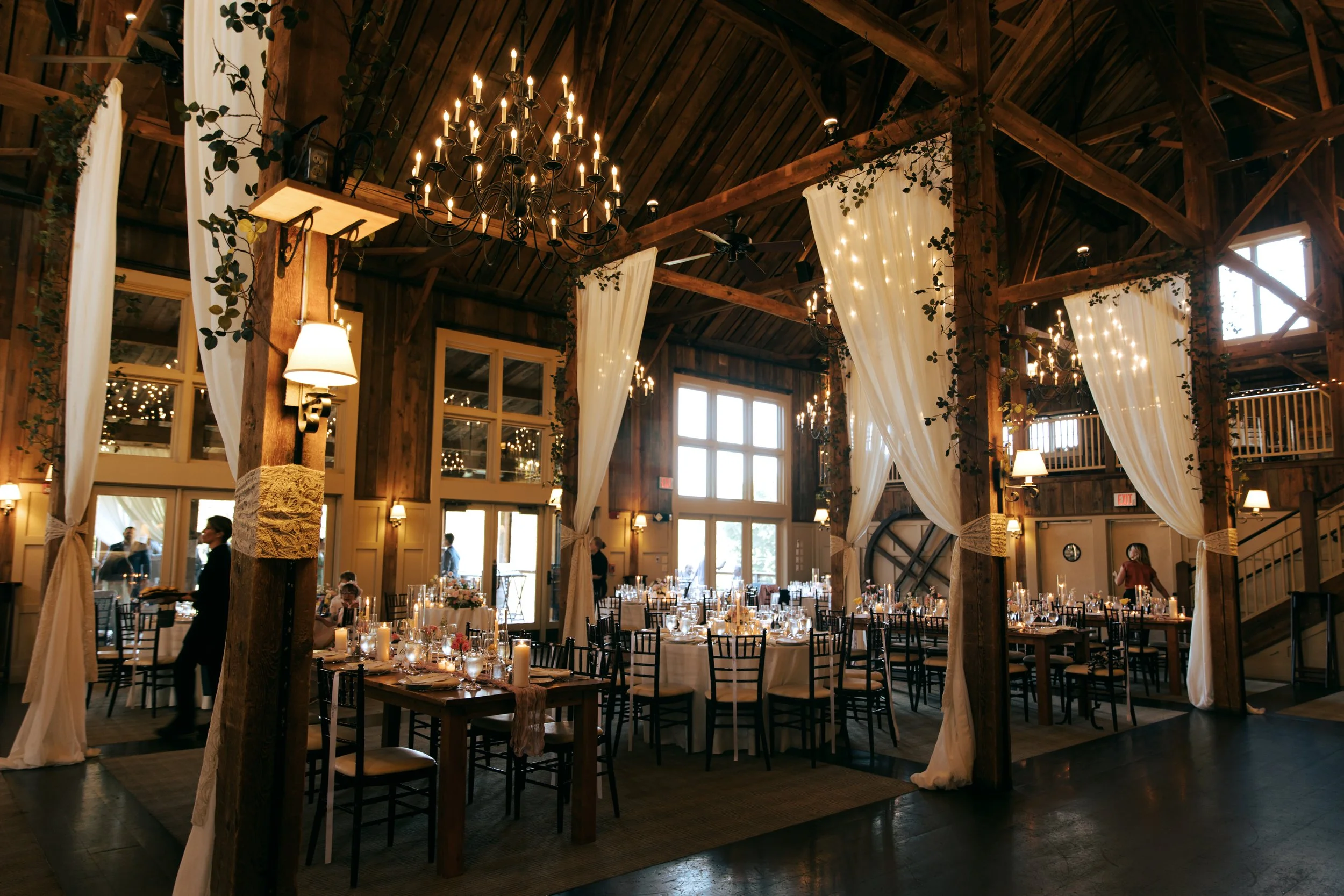 A decorated banquet hall with wooden beams, high ceilings, large windows, chandeliers, and elegant white curtains, set for a wedding reception with tables, candles, and chairs.