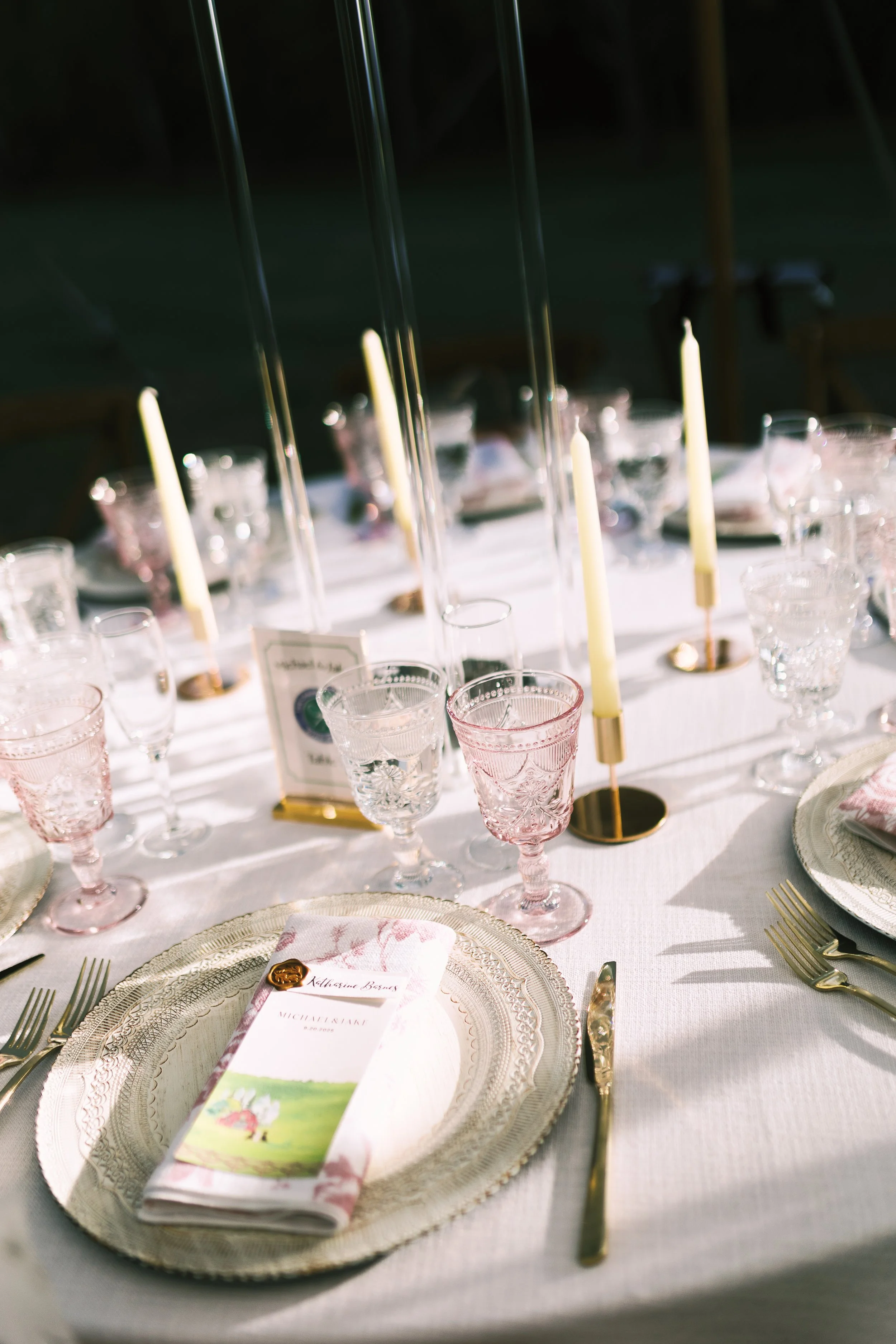 Elegant table setting with gold-rimmed plates, pink-tinted glassware, gold utensils, a white menu with pink floral design, tall candles in gold holders, and a card with a wax seal, set outdoors with natural light.