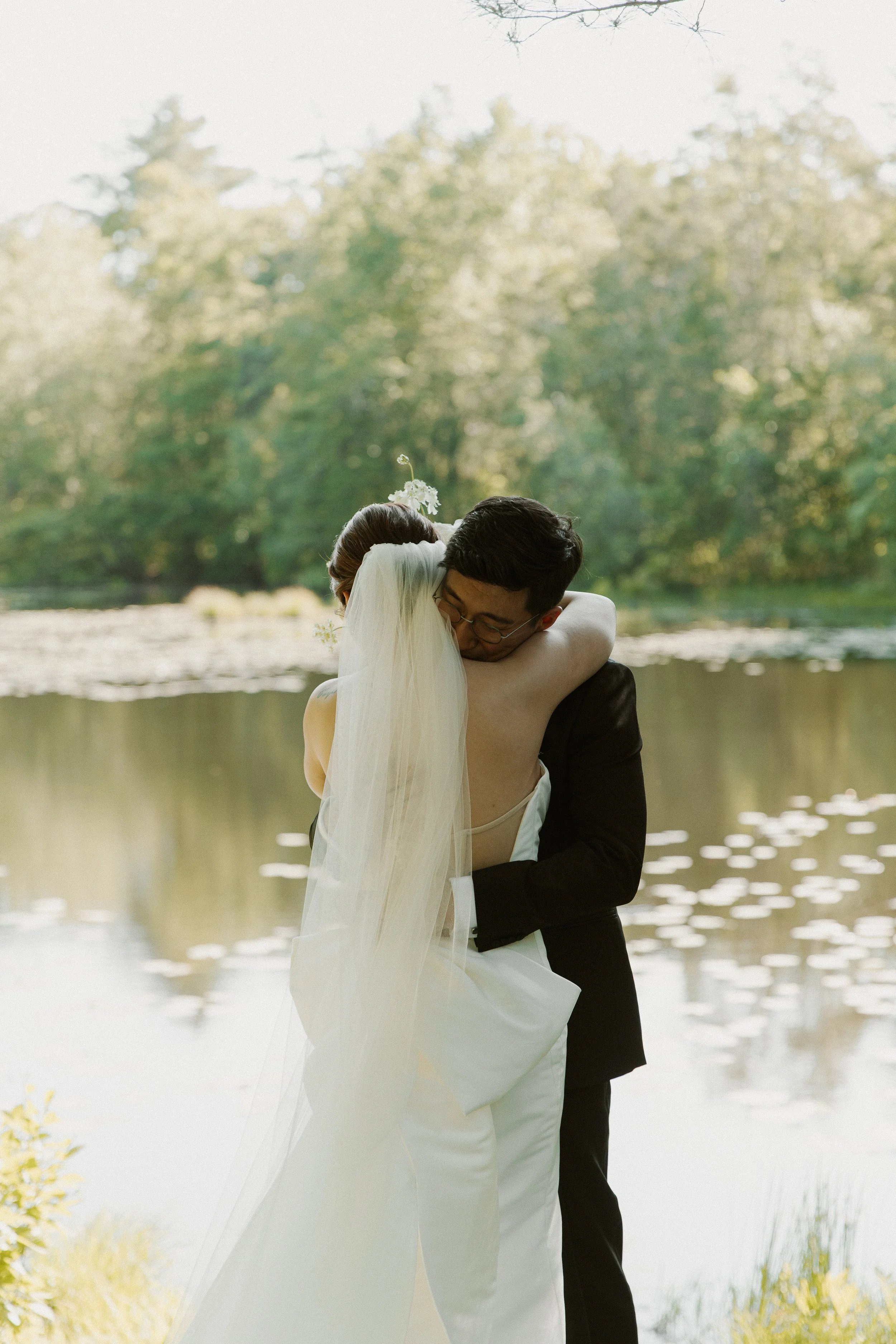 A bride and groom embracing by a pond with trees in the background.