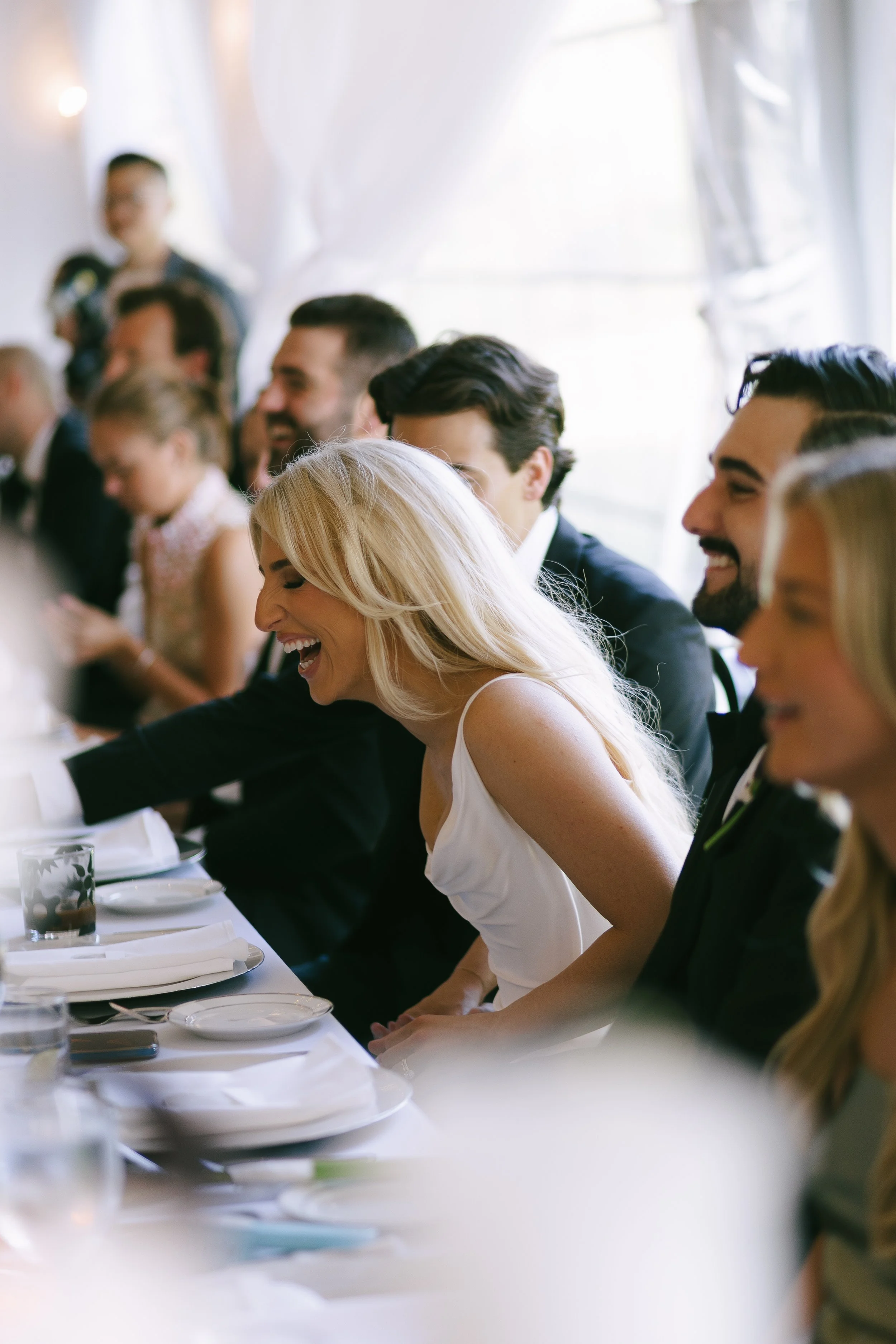 People at a wedding reception are sitting at a long table, laughing and smiling, dressed in formal attire with a blonde woman in a white dress in the foreground.
