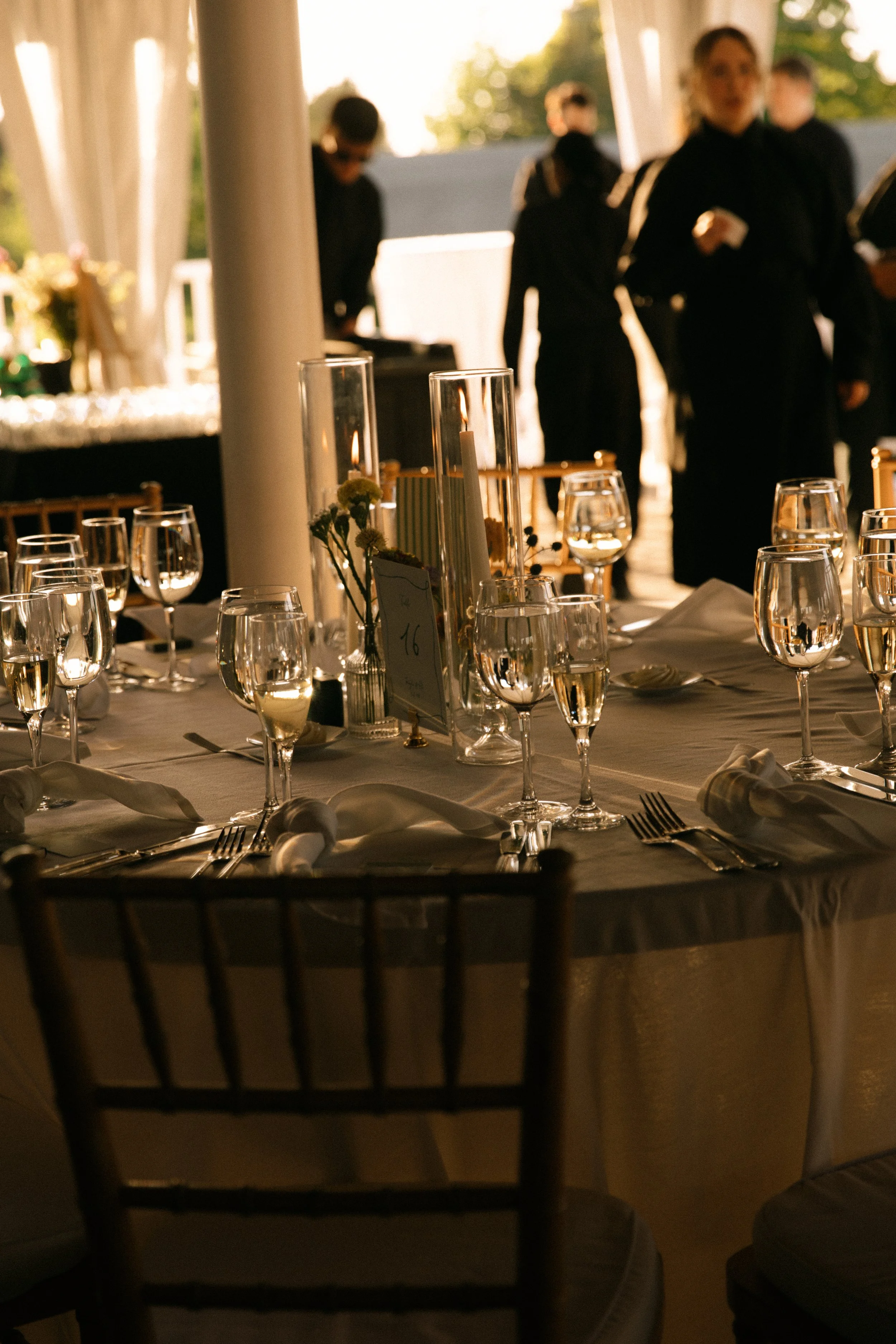 Elegant event table with glasses of champagne and candles, with waitstaff in black uniforms serving in the background at an outdoor setting.