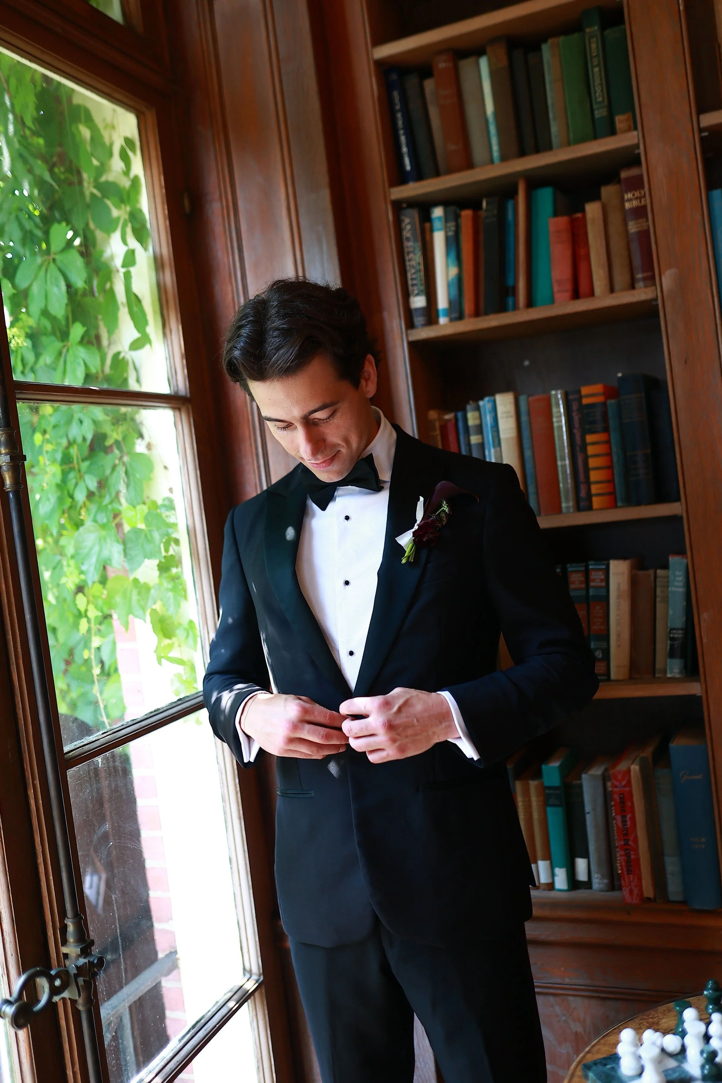 A man in a black tuxedo and bow tie, standing near a window with greenery outside, adjusting his jacket inside a wooden-furnished room with a bookshelf filled with books.