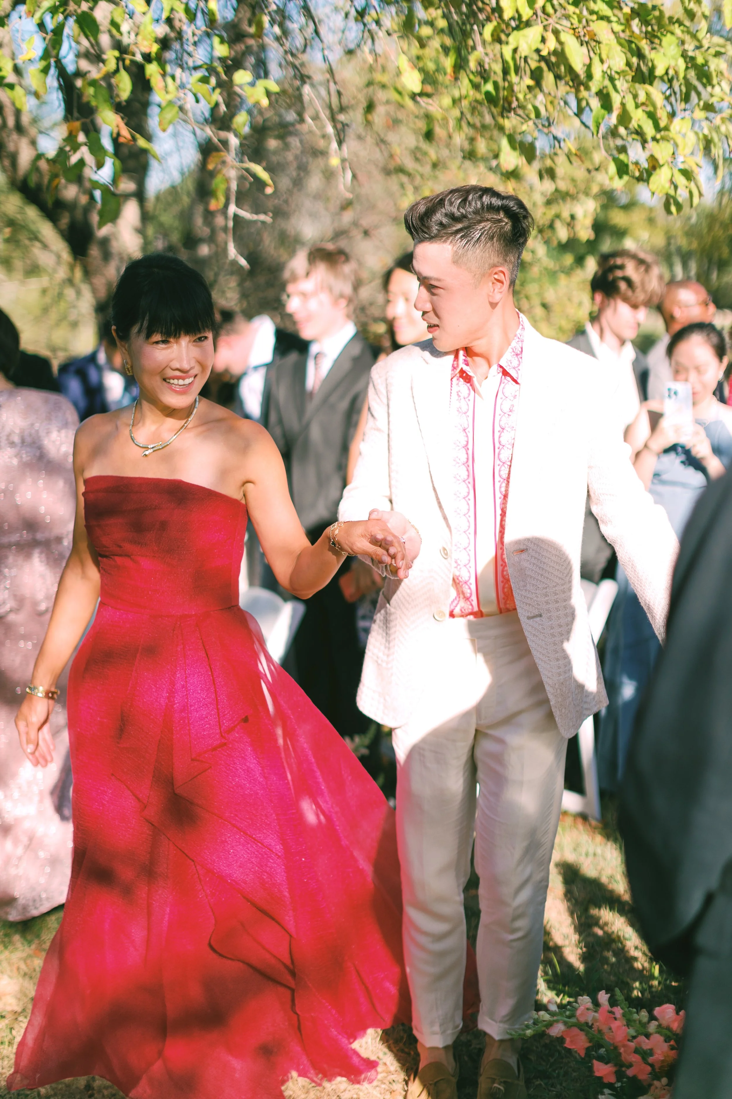 A woman in a strapless red dress dancing with a man in a white suit outdoors during daytime surrounded by a group of people.