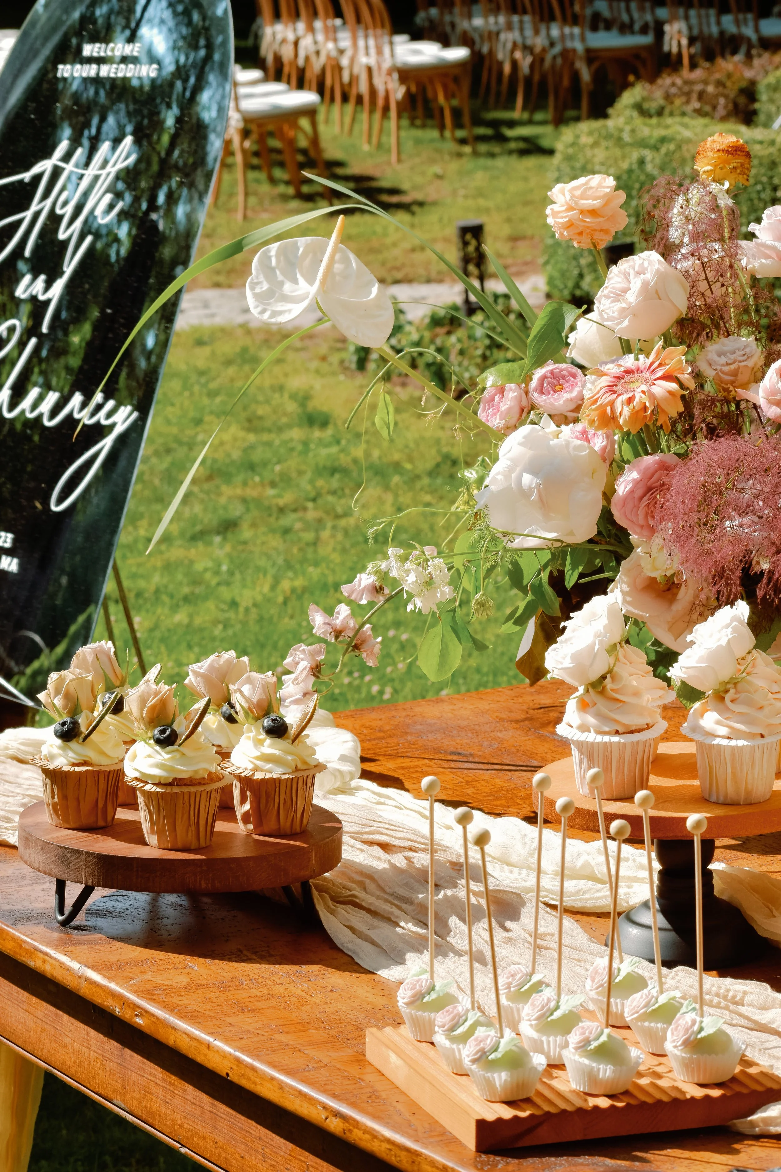 Table with cupcakes and a large floral arrangement at an outdoor wedding reception featuring a welcome sign.