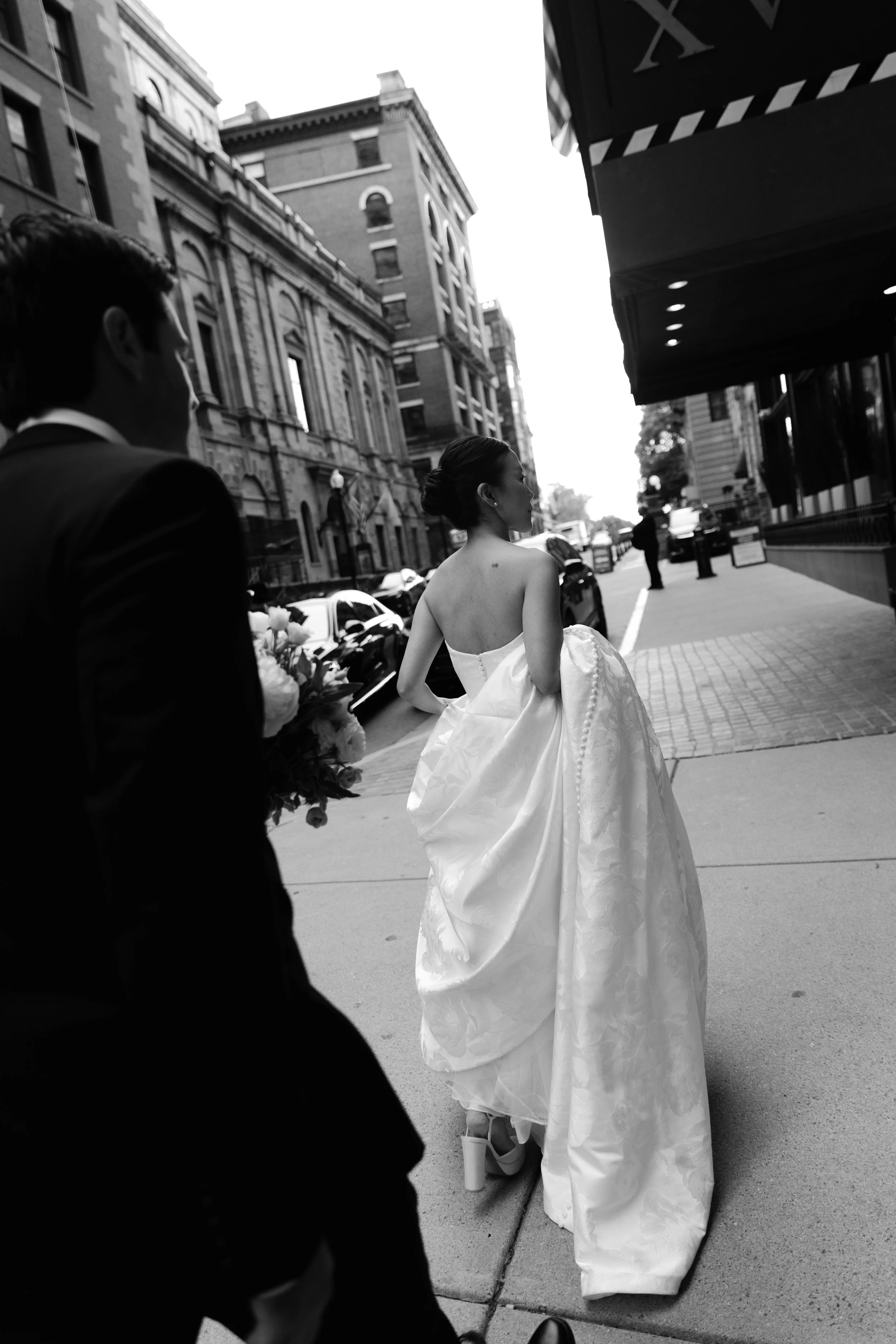 A bride in a strapless wedding gown with a full skirt, walking on a city sidewalk, with a groom holding a bouquet behind her, during daytime.