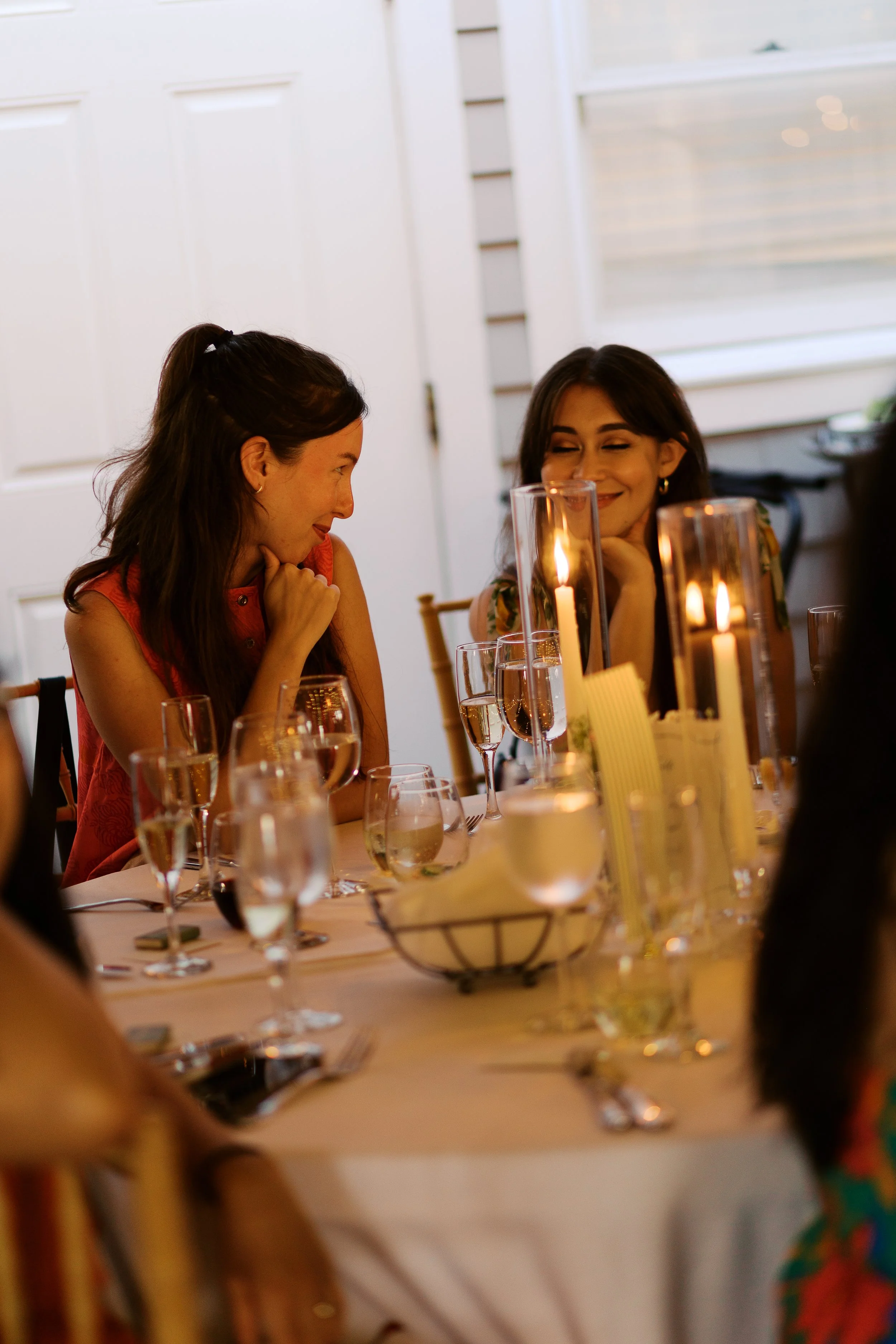 Two women smiling and chatting at a dinner table with lit candles and wine glasses.