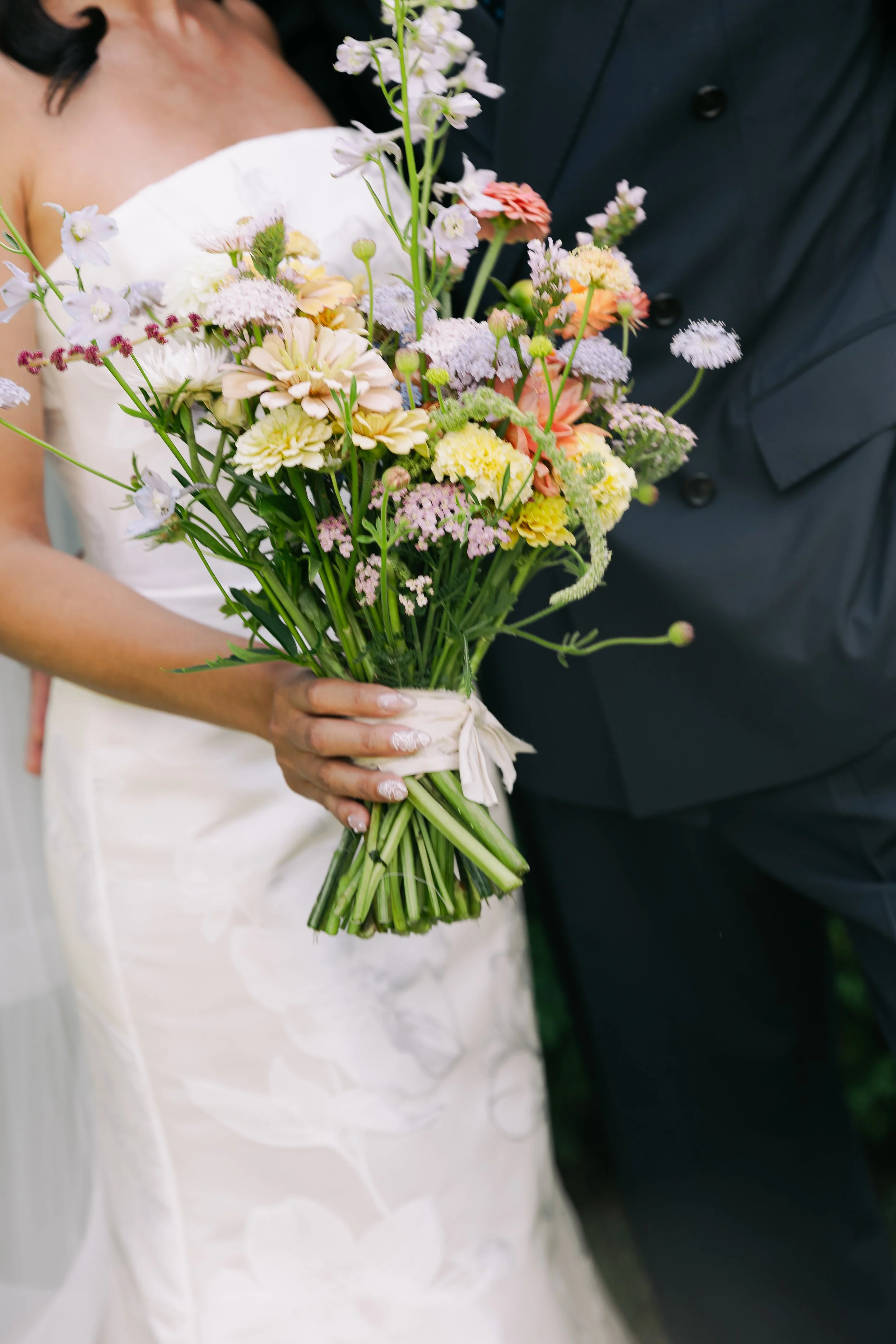 A bride holding a bouquet of mixed flowers, blended with light colors, with a groom standing beside her. The bride's wedding dress and the groom's tuxedo are visible.