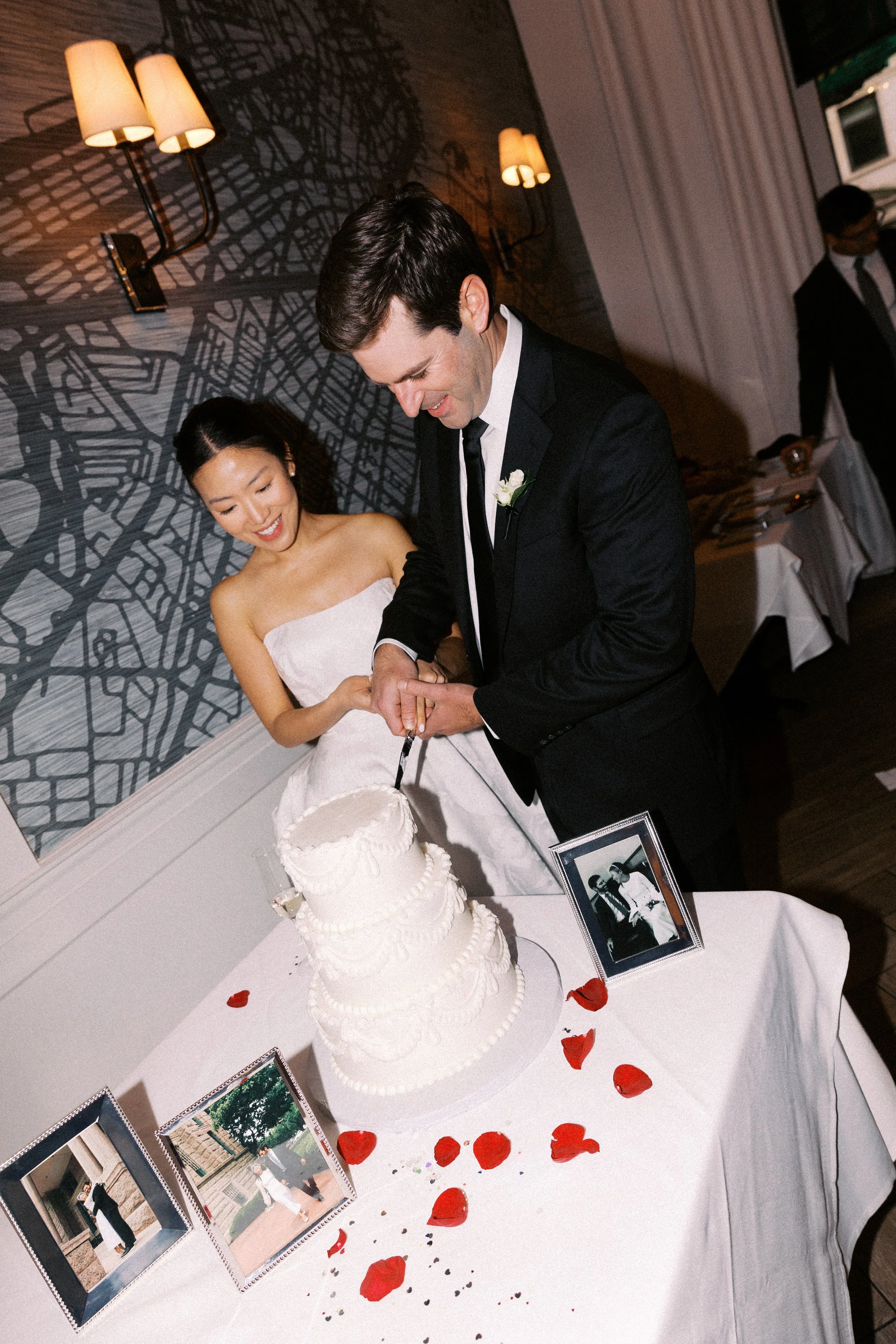 A newlywed couple cutting a wedding cake at their reception. The bride is smiling, wearing a strapless white wedding dress, and the groom is dressed in a black tuxedo. The table is decorated with red rose petals and framed photos. The background incl