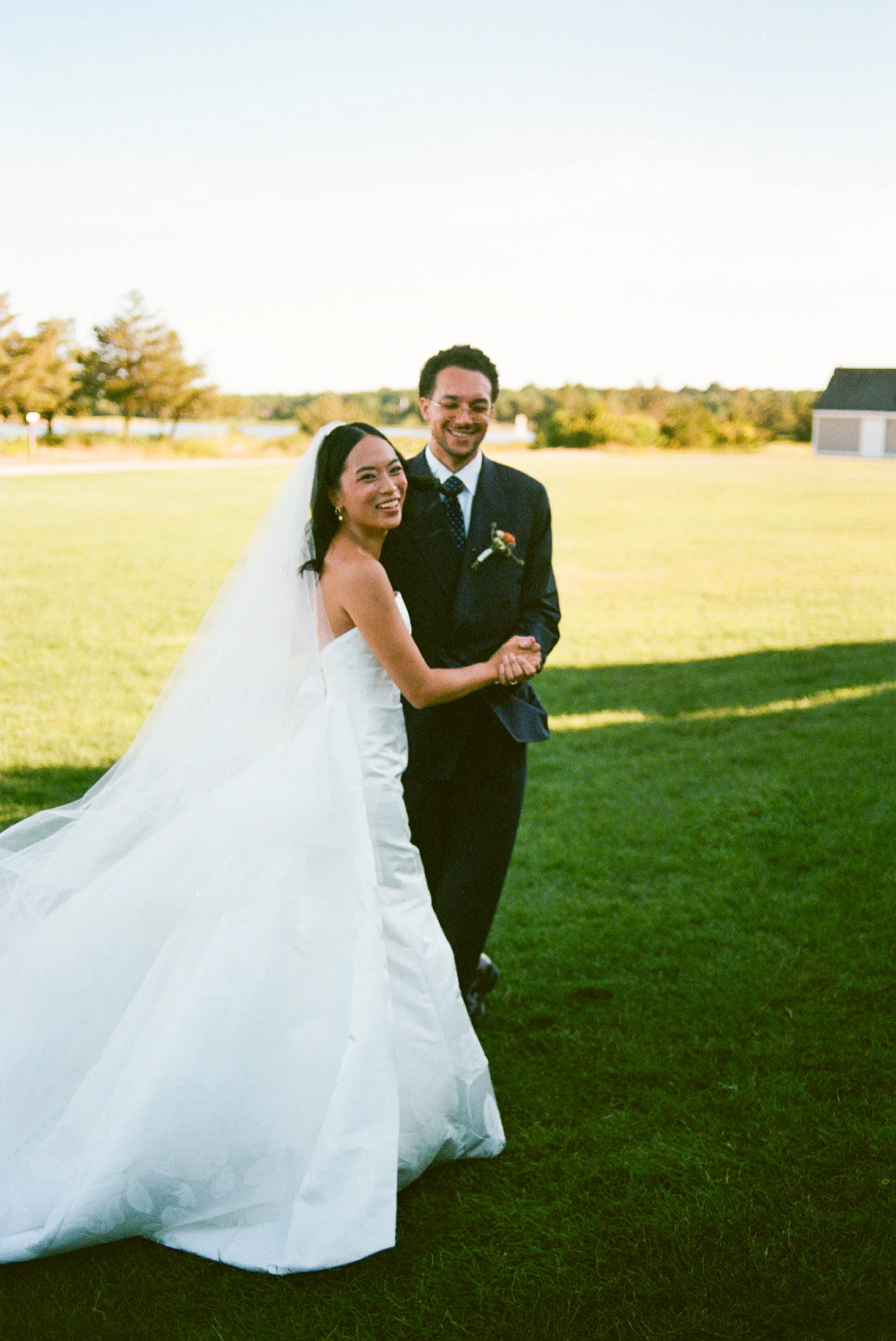 A bride and groom smiling and holding hands outdoors on their wedding day, standing on a grassy field with trees and a building in the background under a clear sky.