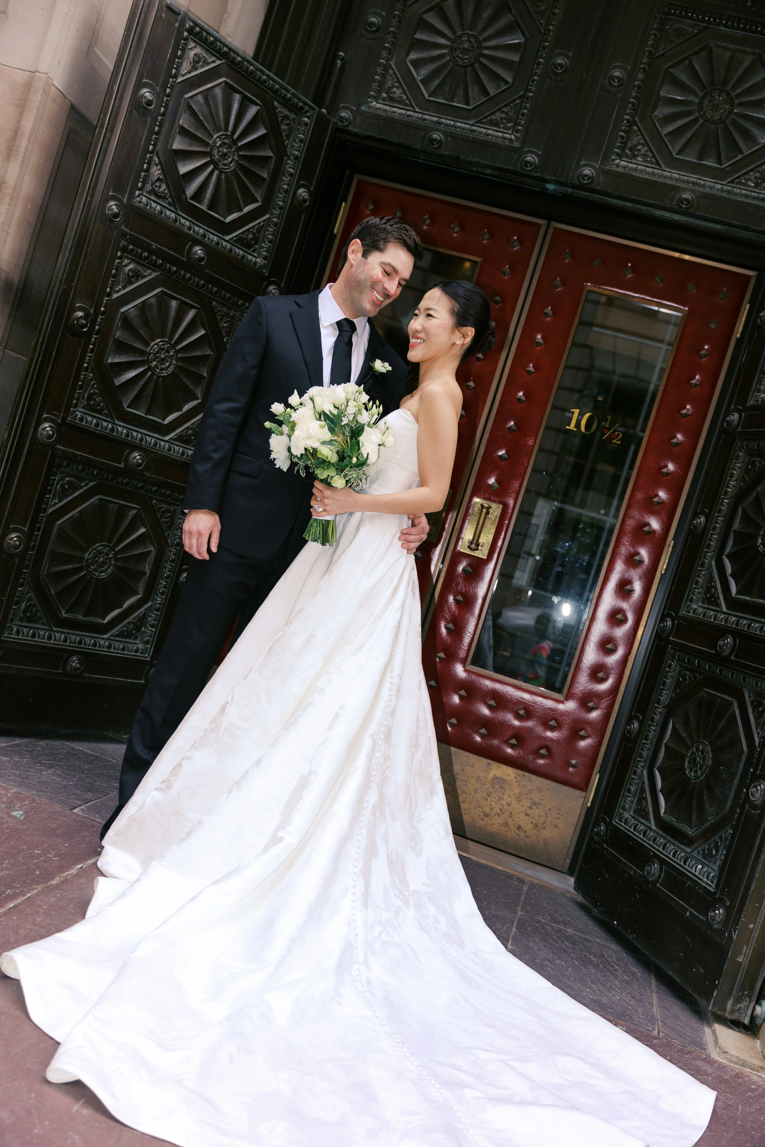 A newlywed couple standing in front of an ornate door, sharing a joyful moment. The bride is holding a bouquet of white flowers, and is wearing a strapless white wedding gown with a long train. The groom is in a dark suit, smiling at her.
