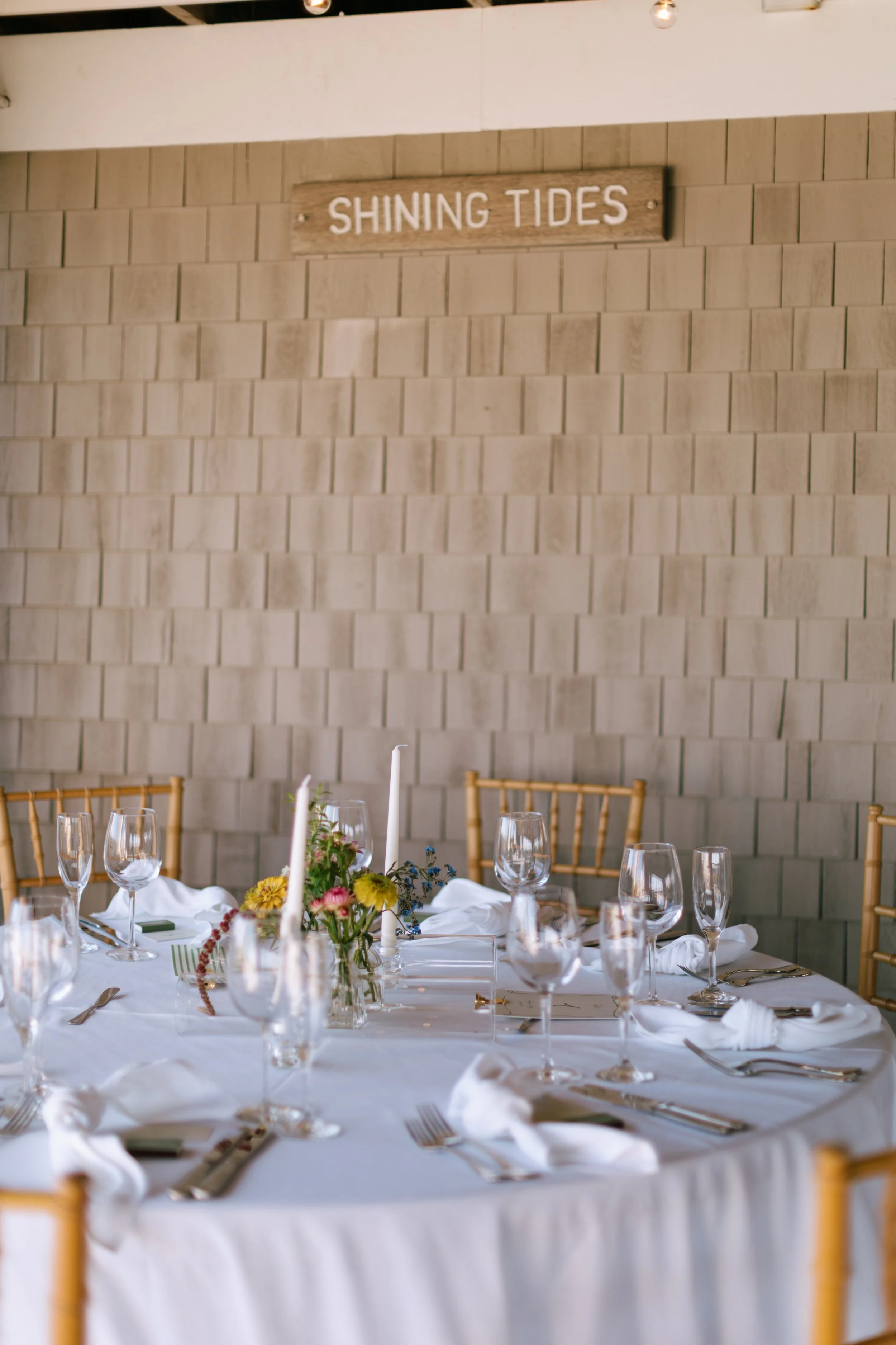 A round dining table set for a formal event with white tablecloths, glassware, silverware, and folded napkins, featuring a centerpiece of mixed flowers and taper candles. There are wooden chairs with vertical slats around the table, and a wooden sign