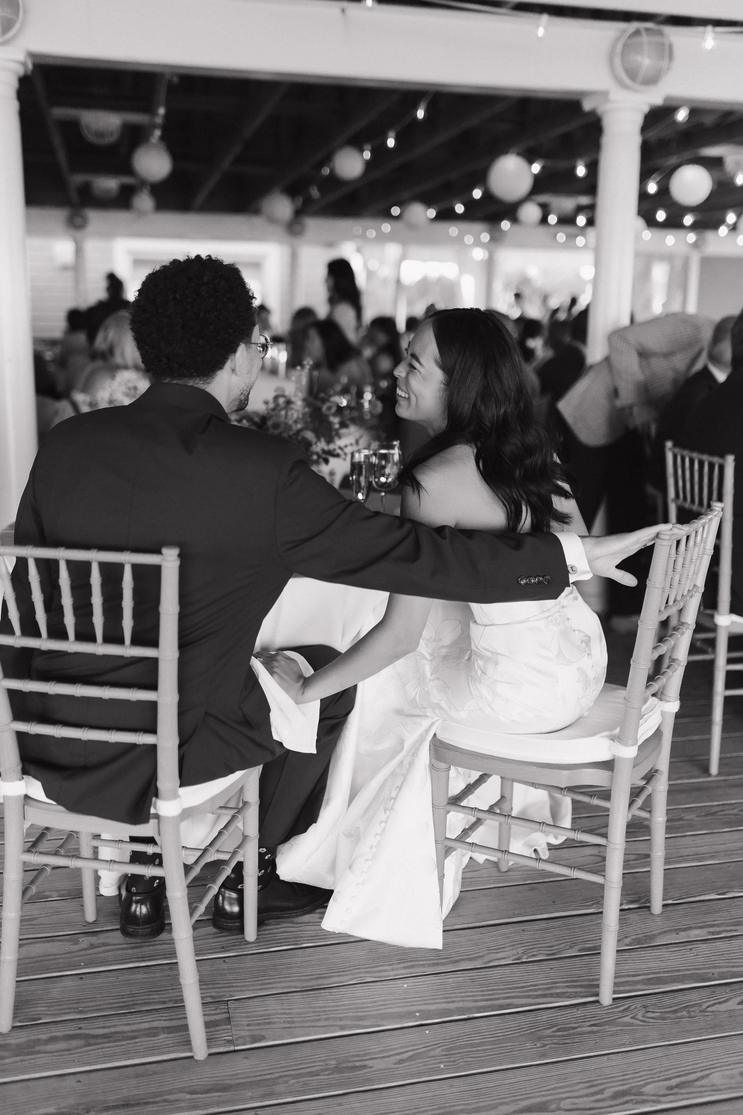 A black and white photo of a man and woman sitting at a table, smiling and holding hands. They are at a wedding reception with many guests in the background, decorated with string lights and paper lanterns.