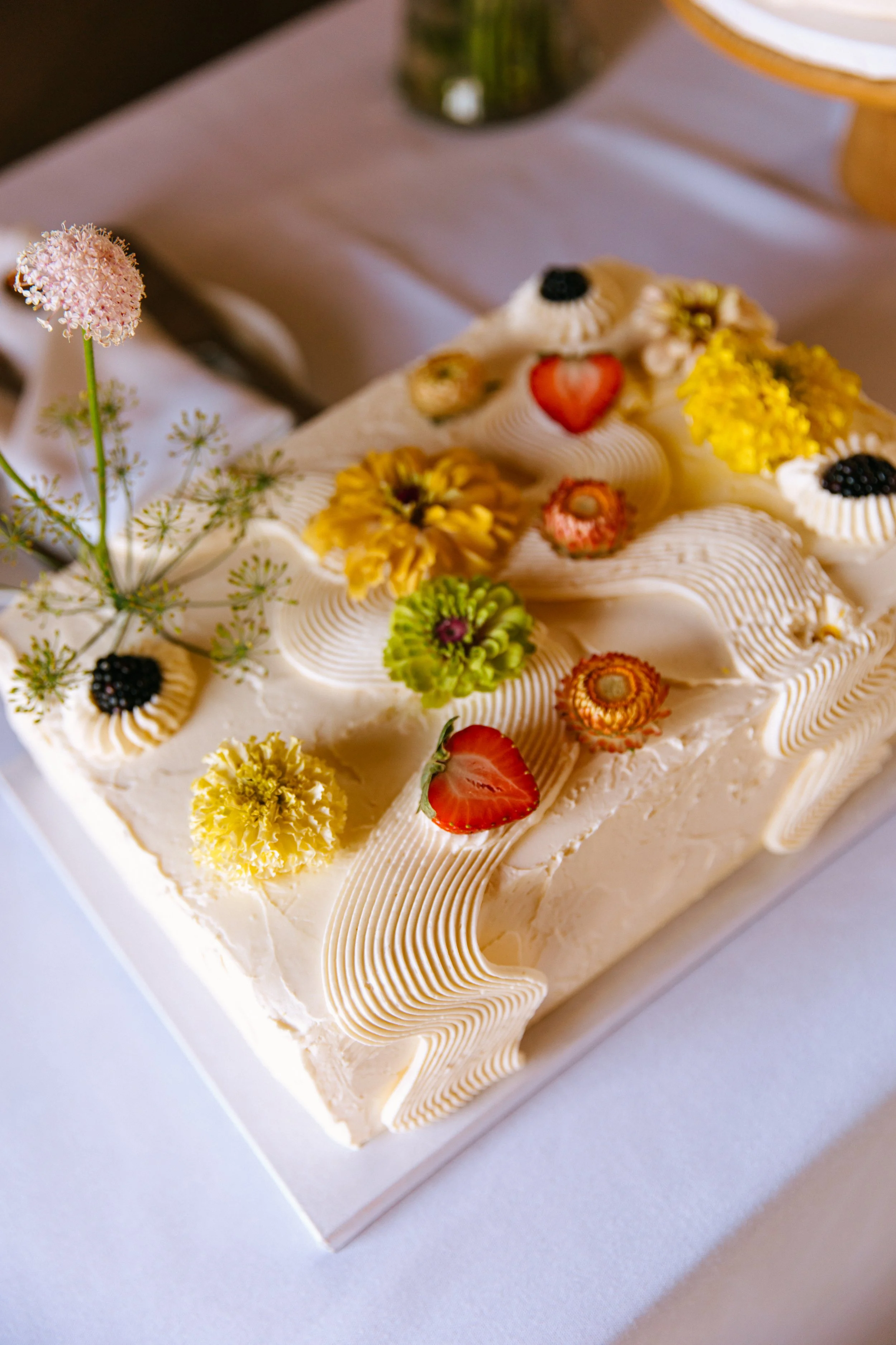 Decorative cake with white icing, colorful flowers, strawberries, and blackberries on top, on a white tablecloth.