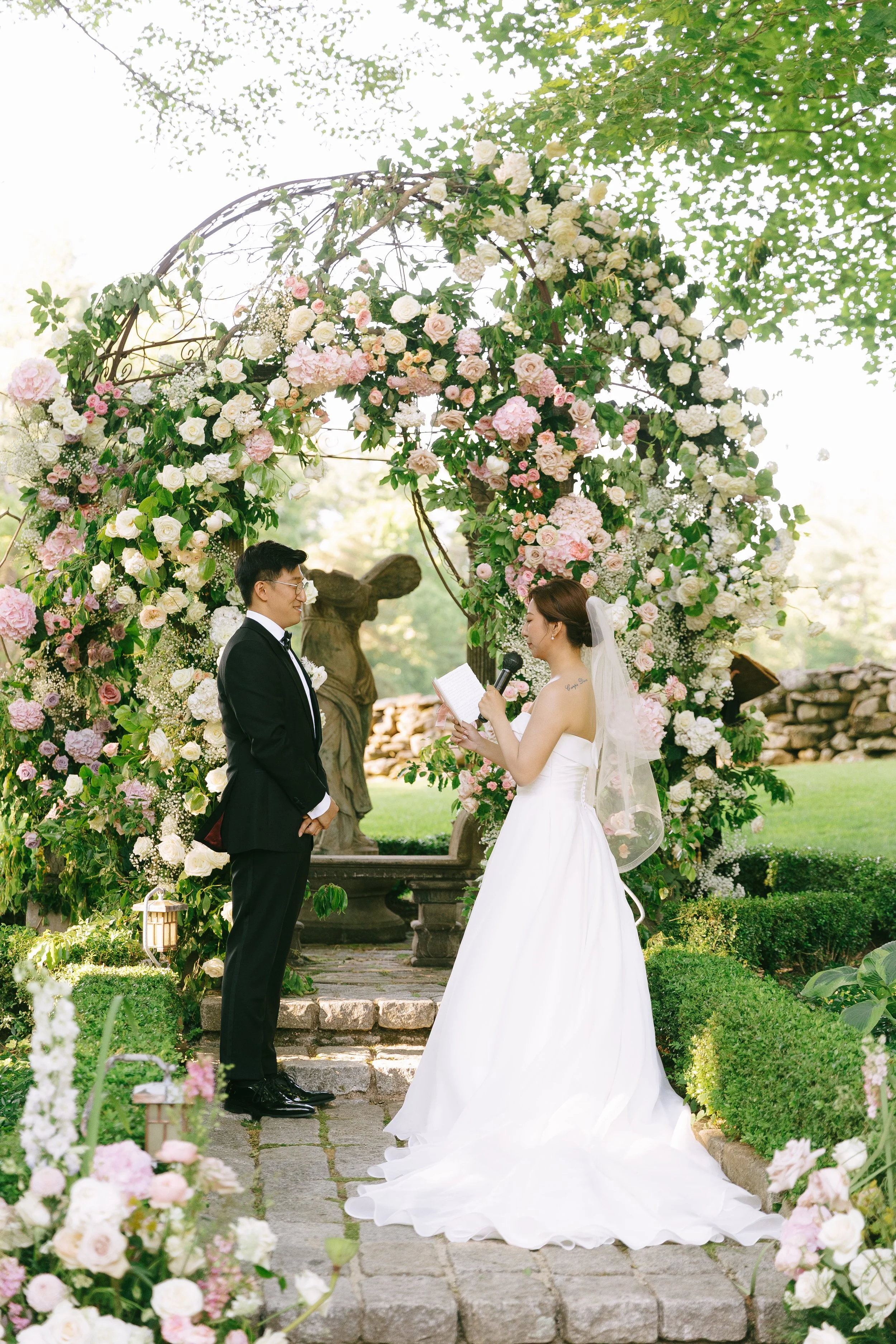 A bride and groom standing under a floral wedding arch during their outdoor wedding ceremony, with a statue in the background.