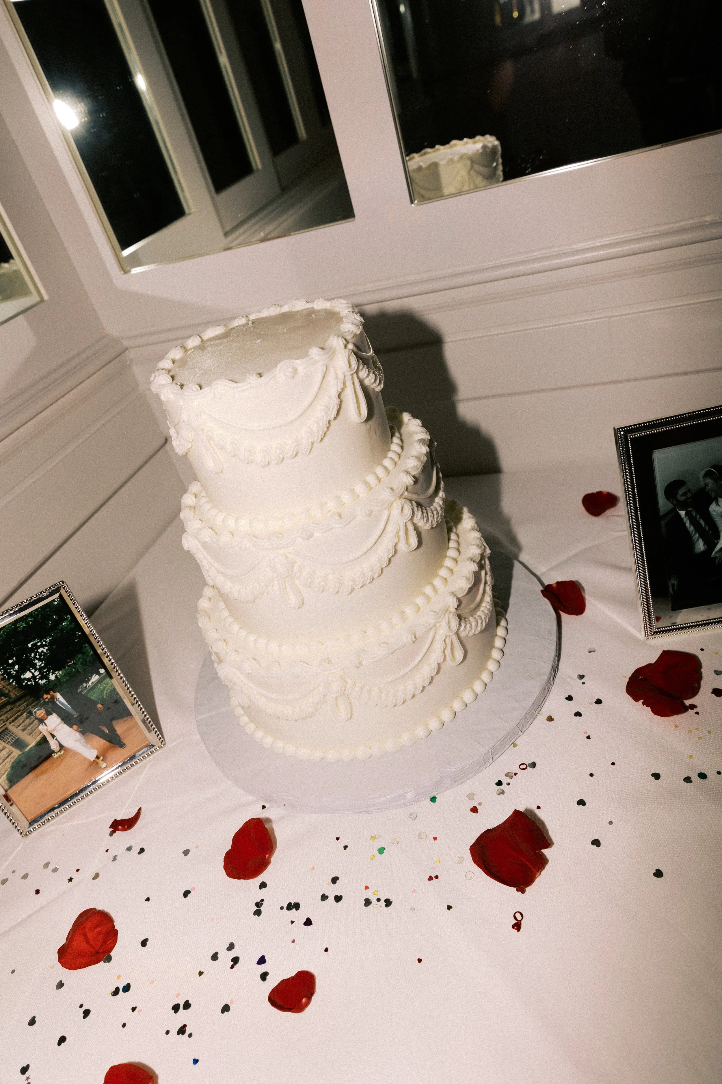 A four-tiered white wedding cake with ornate piping decorations displayed on a table. The table is decorated with red rose petals, scattered confetti, and framed photographs, inside a room with white walls and windows.