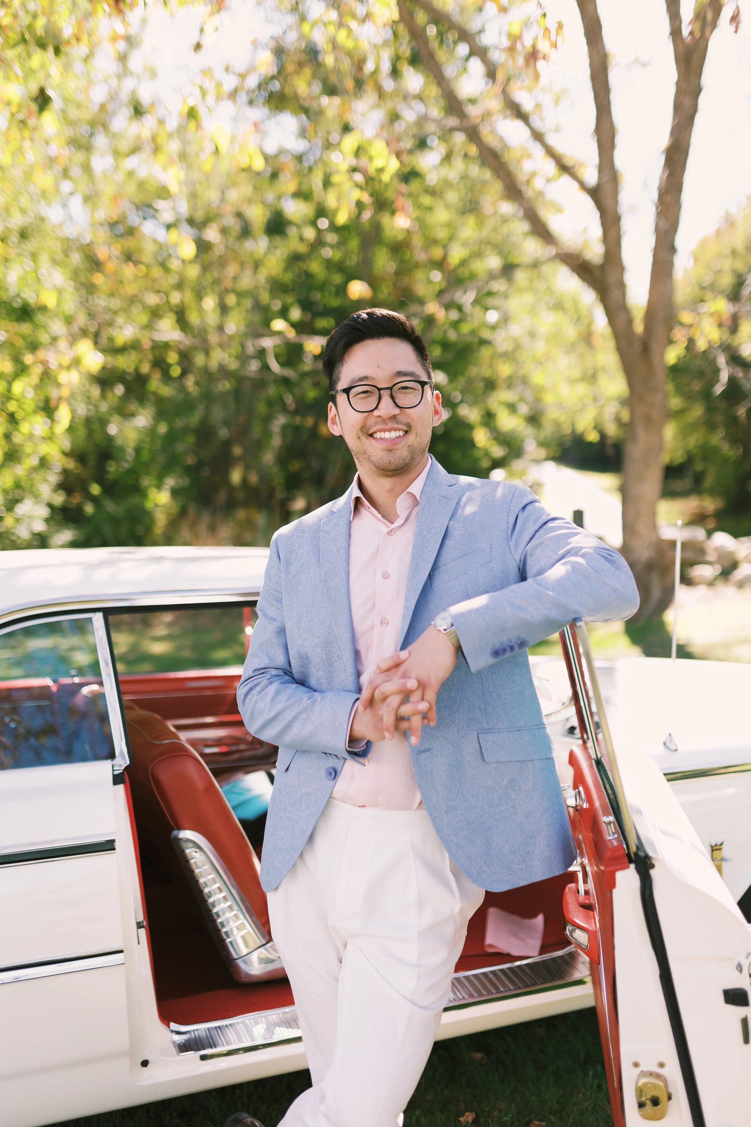 A smiling man with glasses, wearing a light blue blazer and white pants, standing outdoors next to a vintage white car with red interior and open door, on a sunny day with trees and greenery in the background.