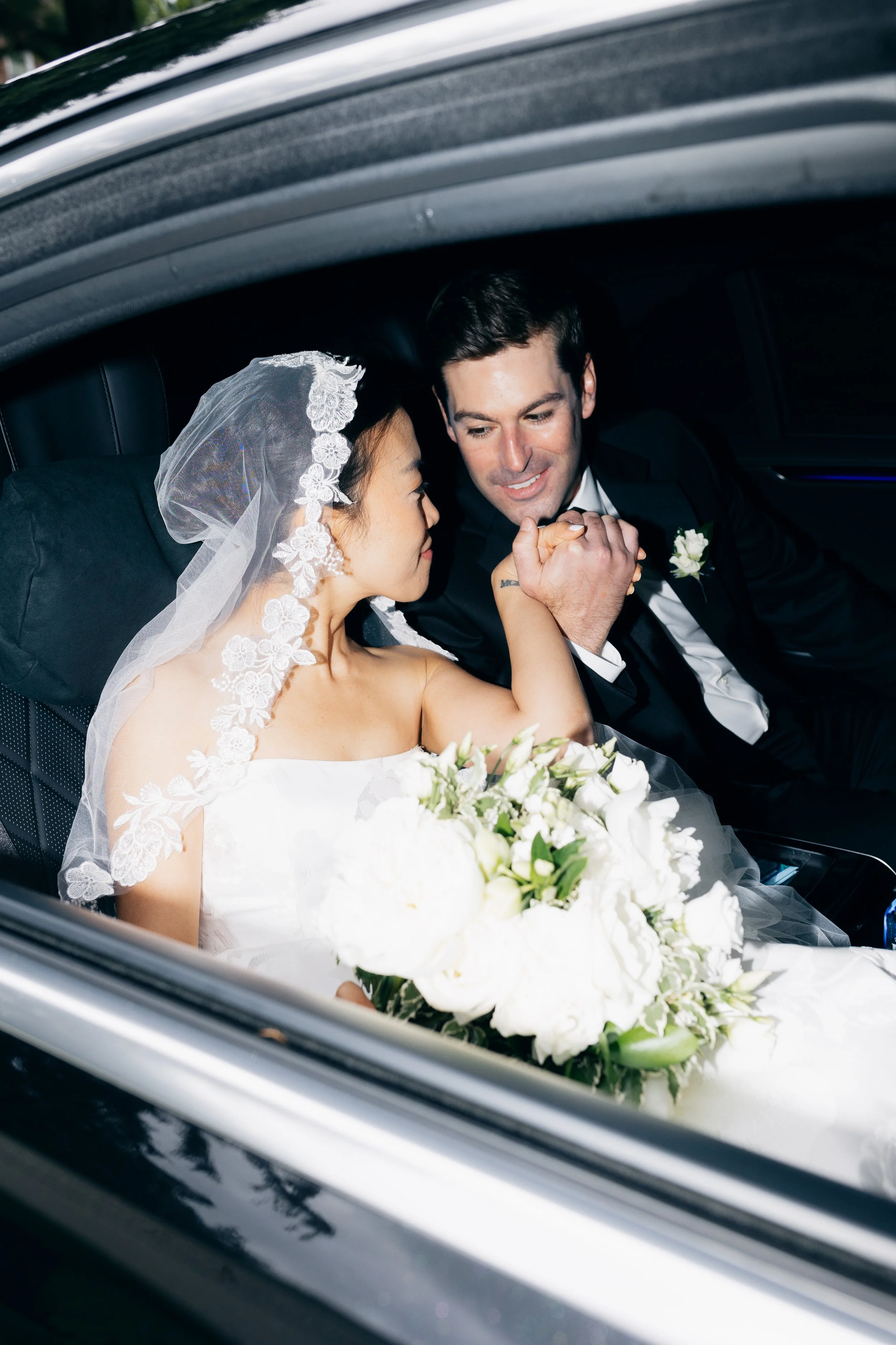 Bride and groom sitting in a car, holding hands, smiling, with the bride wearing a wedding dress, veiled, and holding a bouquet of white flowers.